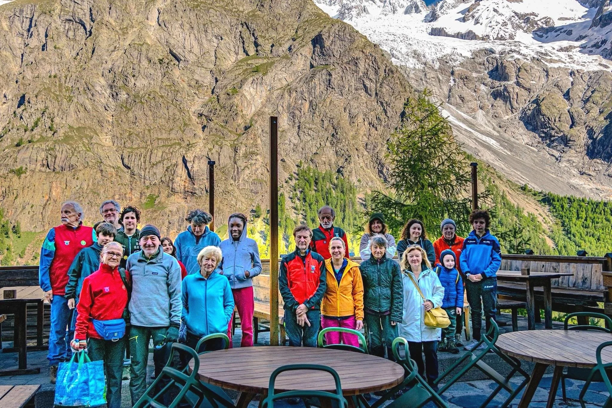 Gruppo di persone che soggiorna all'aperto in montagna con sfondo di pareti di roccia e neve, alcune sedute e altre in piedi, indossando abbigliamento da escursionismo colorato.