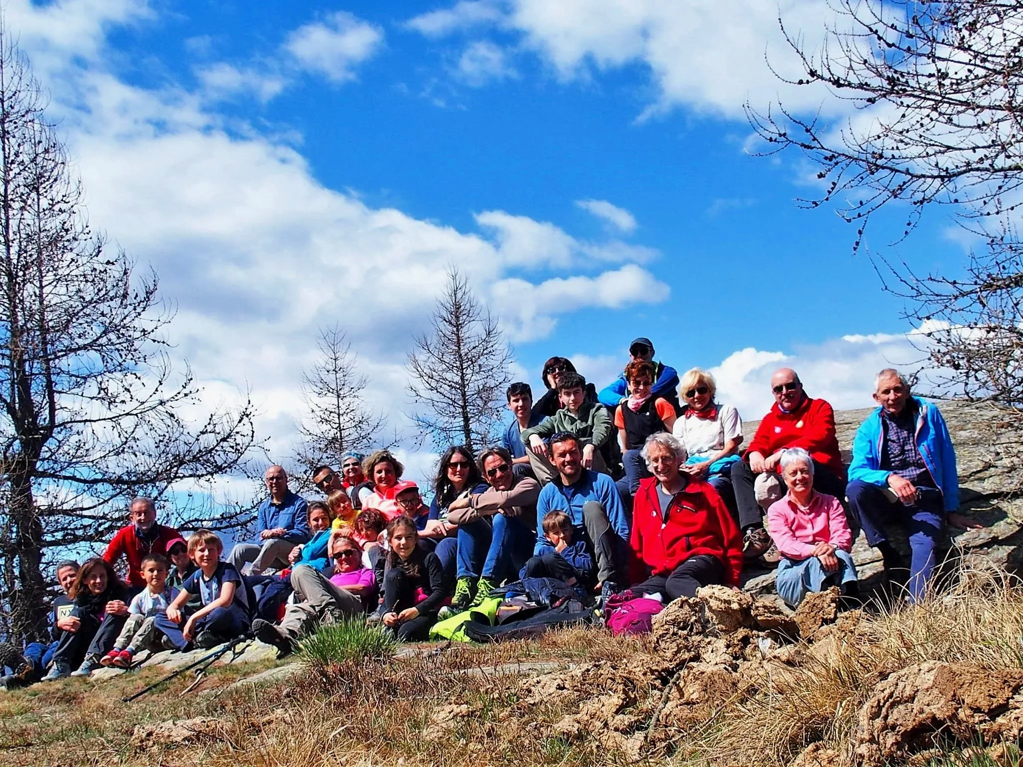 Gruppo di persone, adulti e bambini, seduti su una roccia all'aperto durante una giornata di sole, con cielo azzurro e nuvole. Circondati da alberi e erba secca.