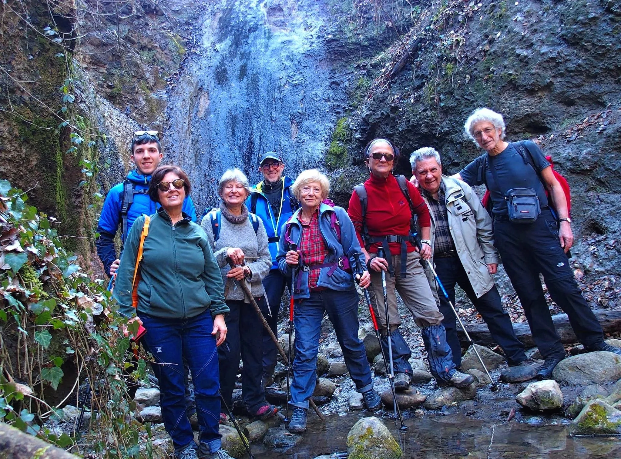 Gruppo di escursionisti in escursione in una zona di montagna con una cascata sullo sfondo, indossando abbigliamento adatto all'outdoor e con bastoni da trekking.