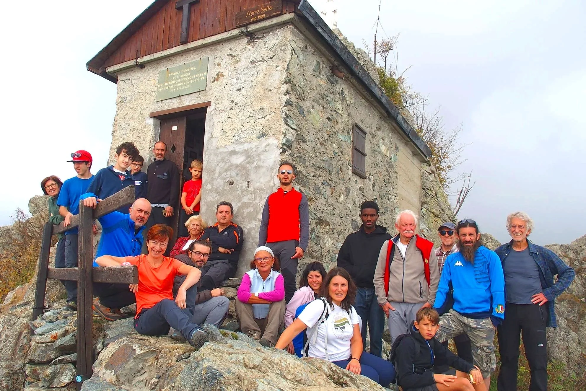 Gruppo di persone che si riuniscono davanti a una piccola casetta di pietra in montagna durante una gita, con alcuni che posano seduti sui sassi e altri in piedi.