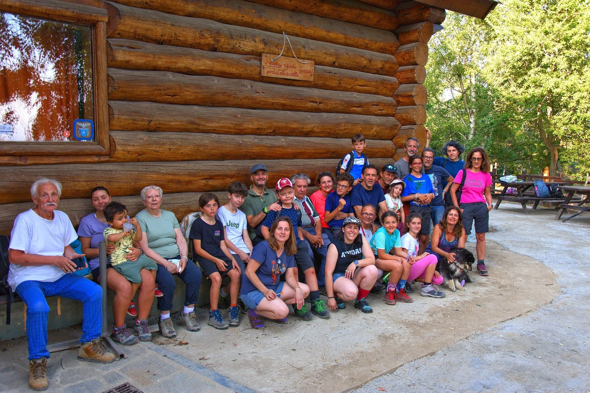 Gruppo di persone di diverse età sorridenti davanti a una baita di legno, alcuni bambini e adulti, in un'area all'aperto alberata.