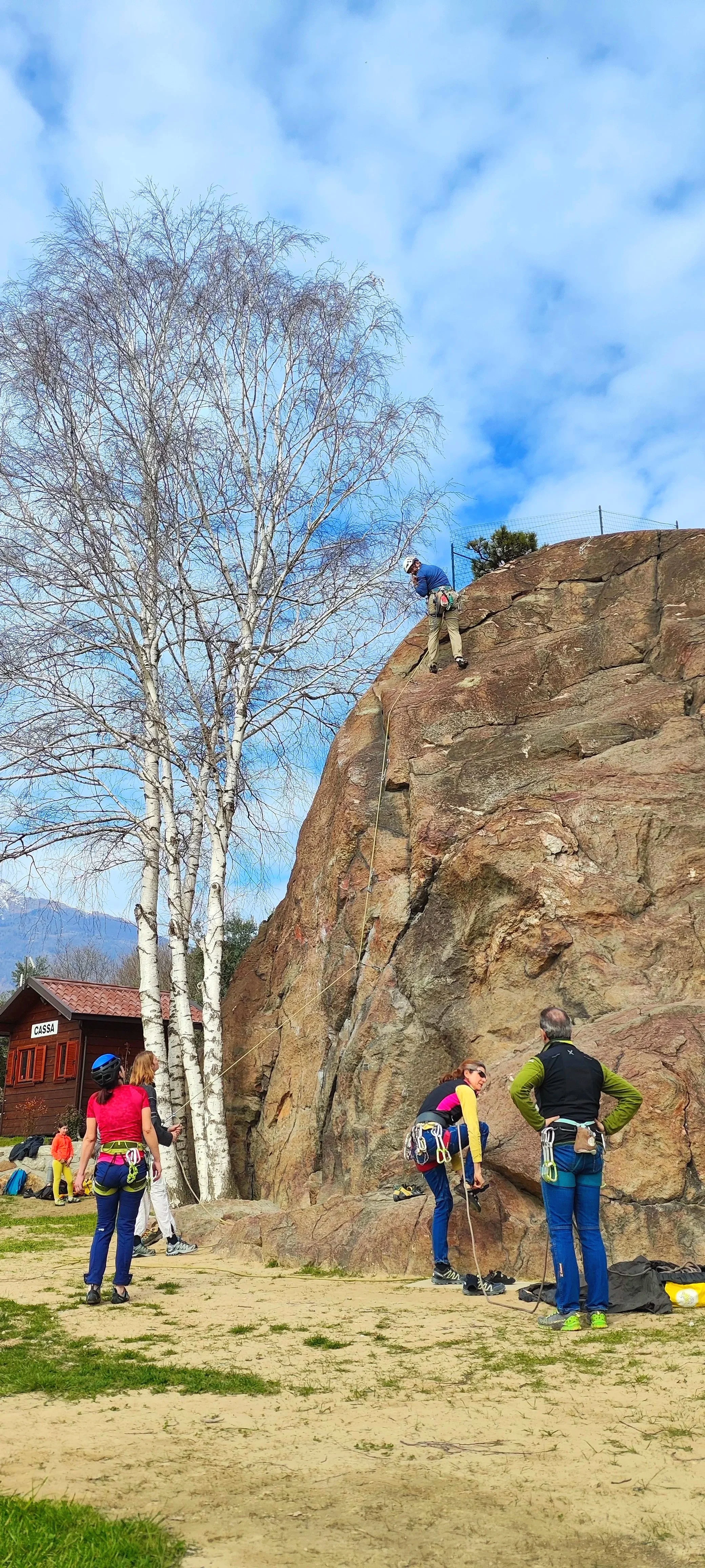 Ragazzi e adulti che praticano l'arrampicata su roccia all'aperto, alcuni in attesa e altri che si allenano sulla parete rocciosa in un'area aperta con un albero senza foglie e un edificio in legno sullo sfondo.