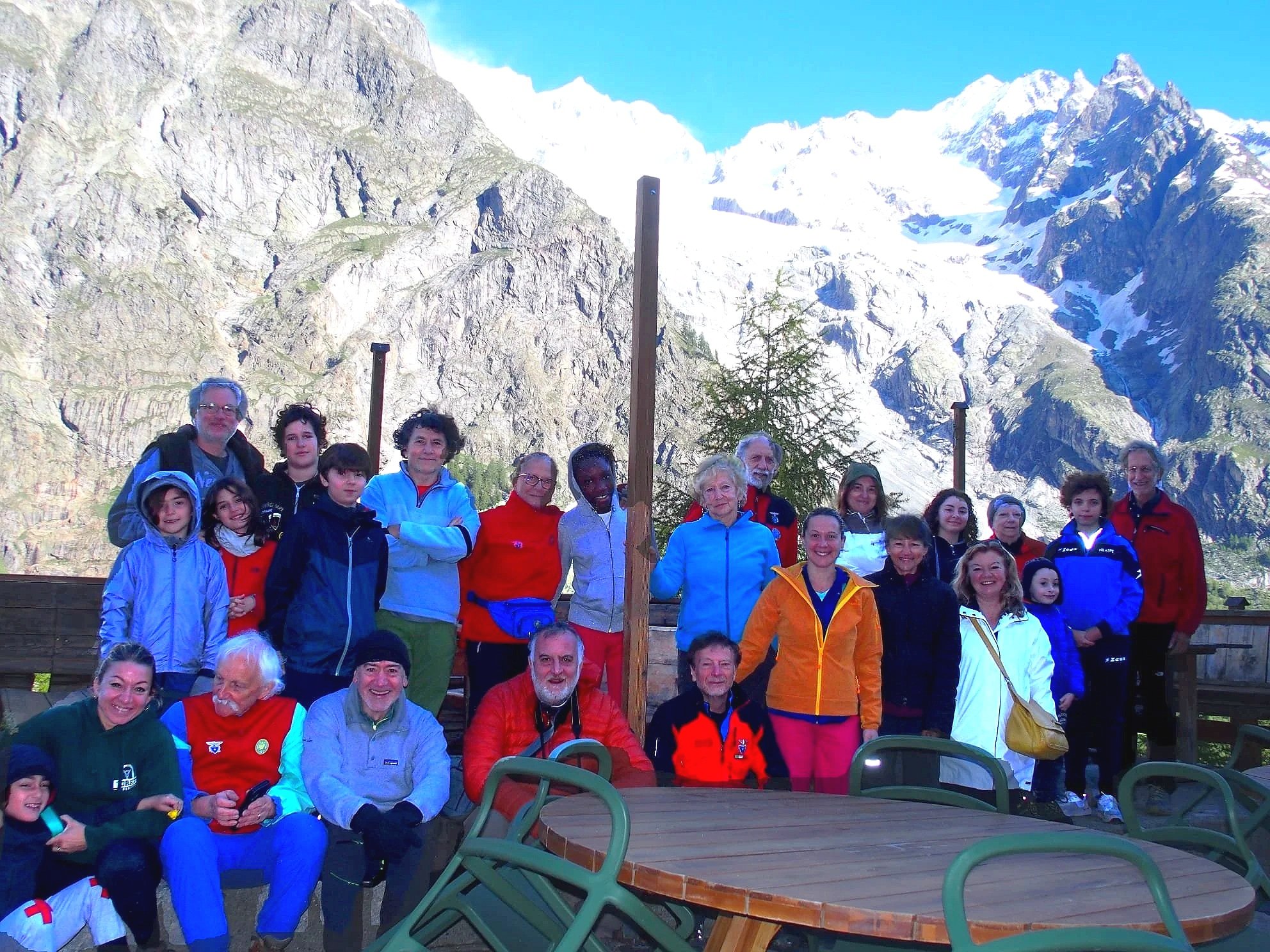 Gruppo di persone in montagna con sfondo di cime innevate e rocce, alcune sedute e altre in piedi, sotto un cielo blu.