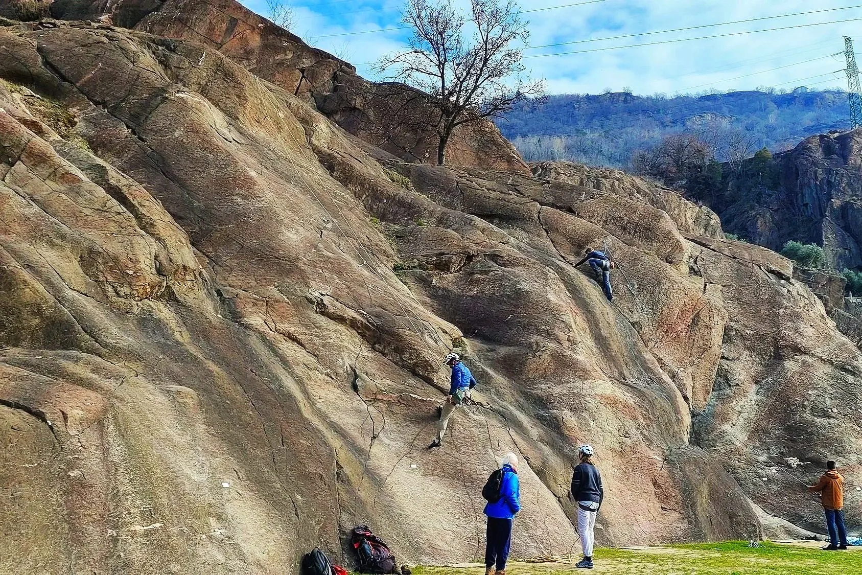 Gruppo di persone che fanno arrampicata su roccia all'aperto in una giornata di sole con cielo blu e alcune montagne sullo sfondo.