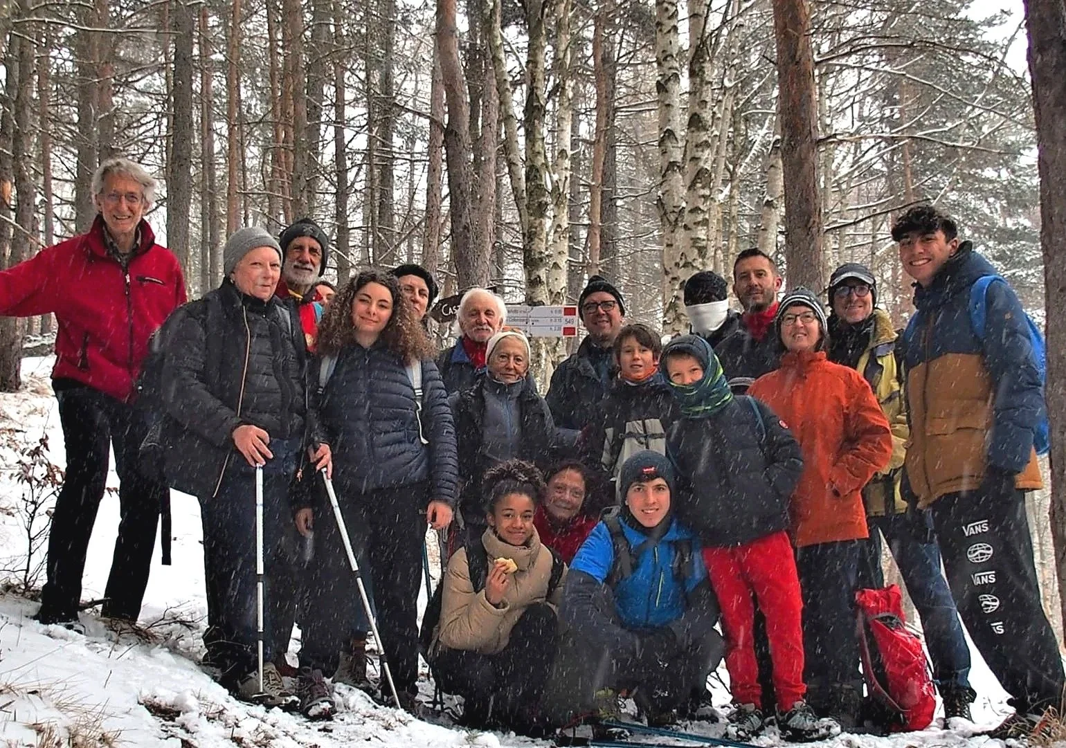 Gruppo di persone in escursione in una foresta innevata, alcune con bastoncini da trekking, tutti vestiti con abbigliamento invernale e sorridenti.