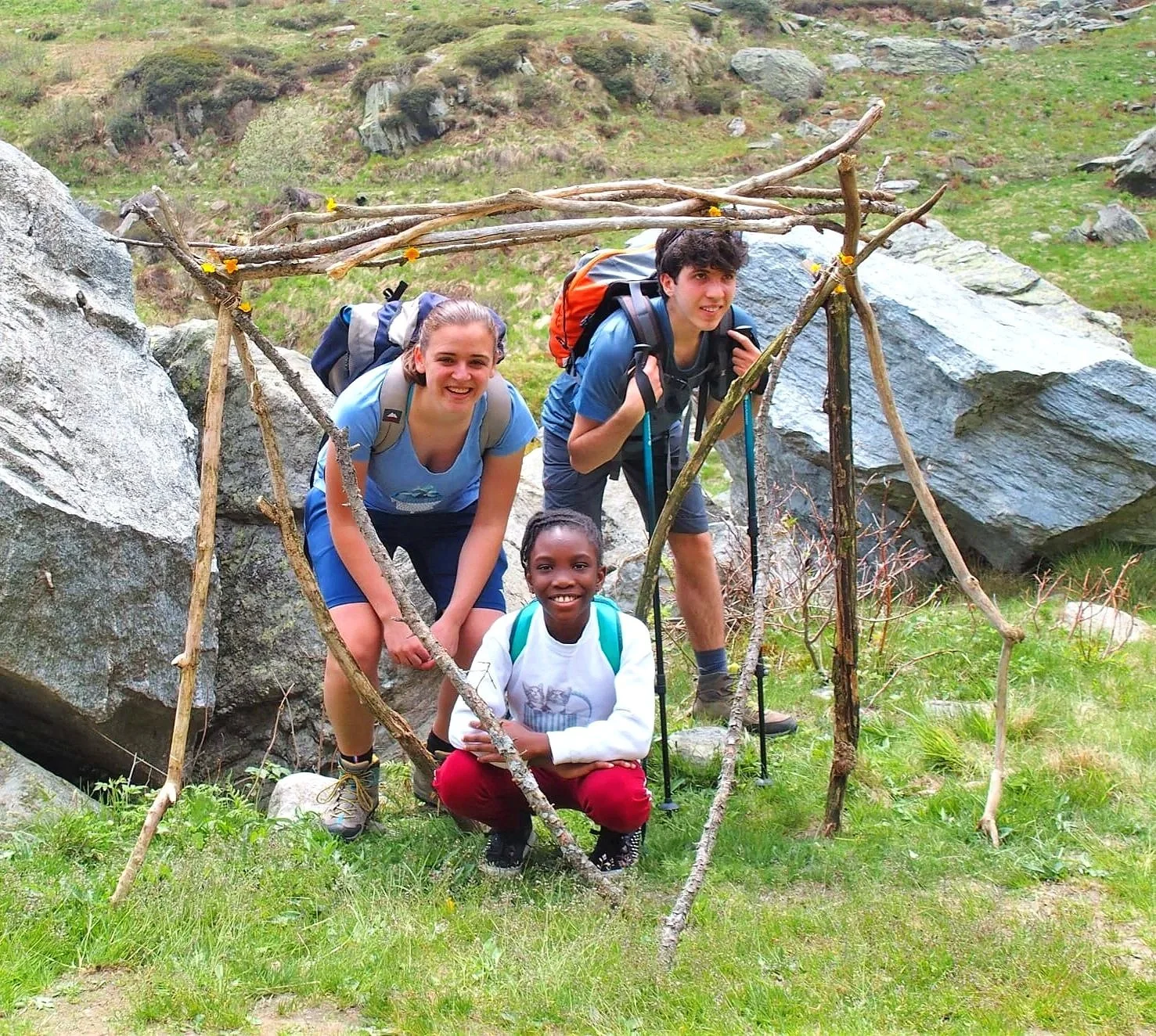Tre giovani all'aperto tra rocce, con una struttura di rami e legnetti decorata con fiori gialli, sorridenti e pronti per un'escursione.