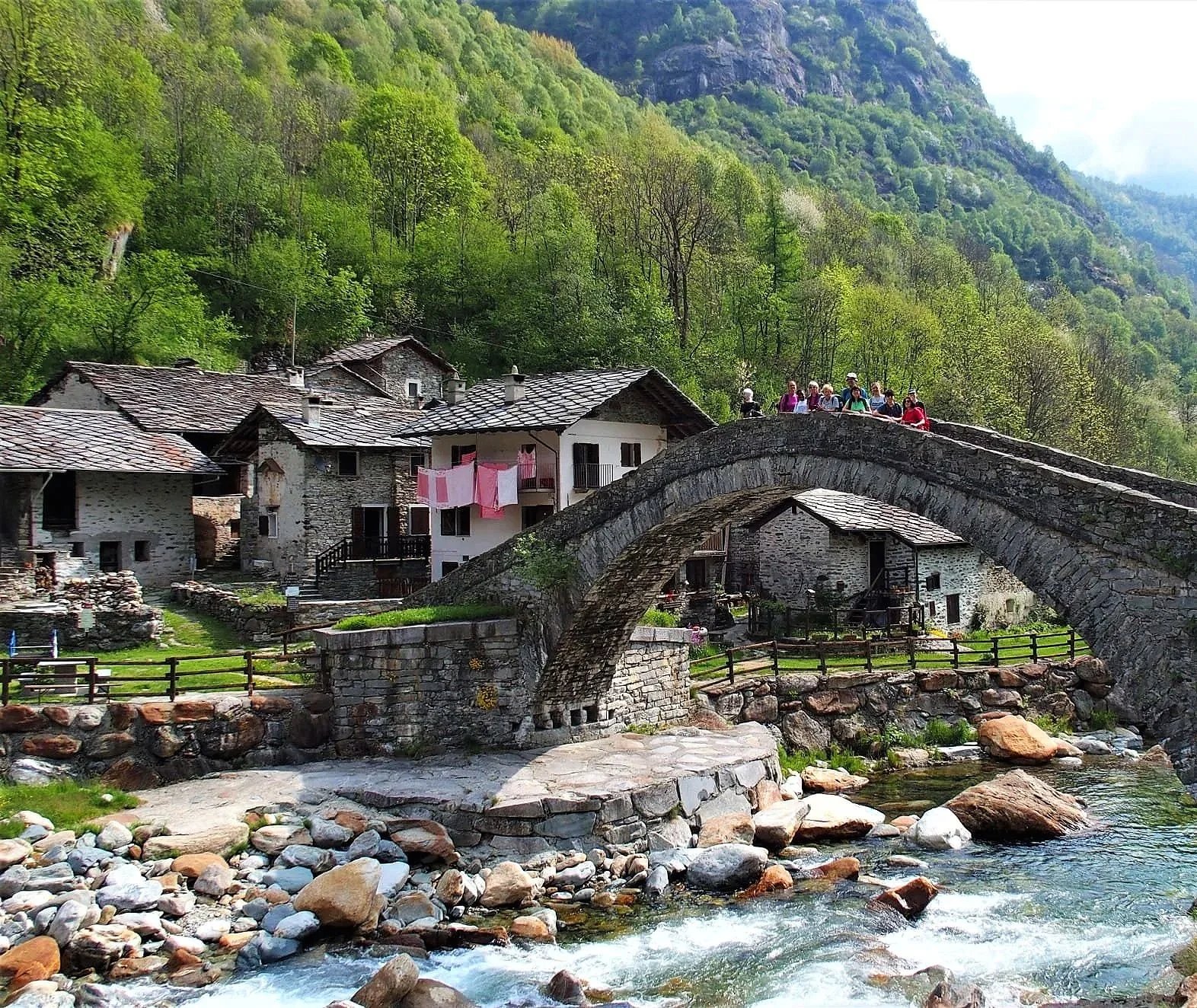 Paesaggio di un piccolo villaggio con case di pietra e un ponte in pietra che attraversa un ruscello, con un gruppo di persone sul ponte, circondato da boschi verdi e colline.