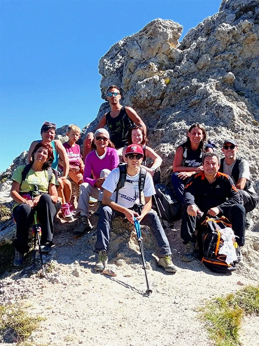 Gruppo di escursionisti con attrezzatura da trekking posano su una salita di montagna con rocce e cielo azzurro.