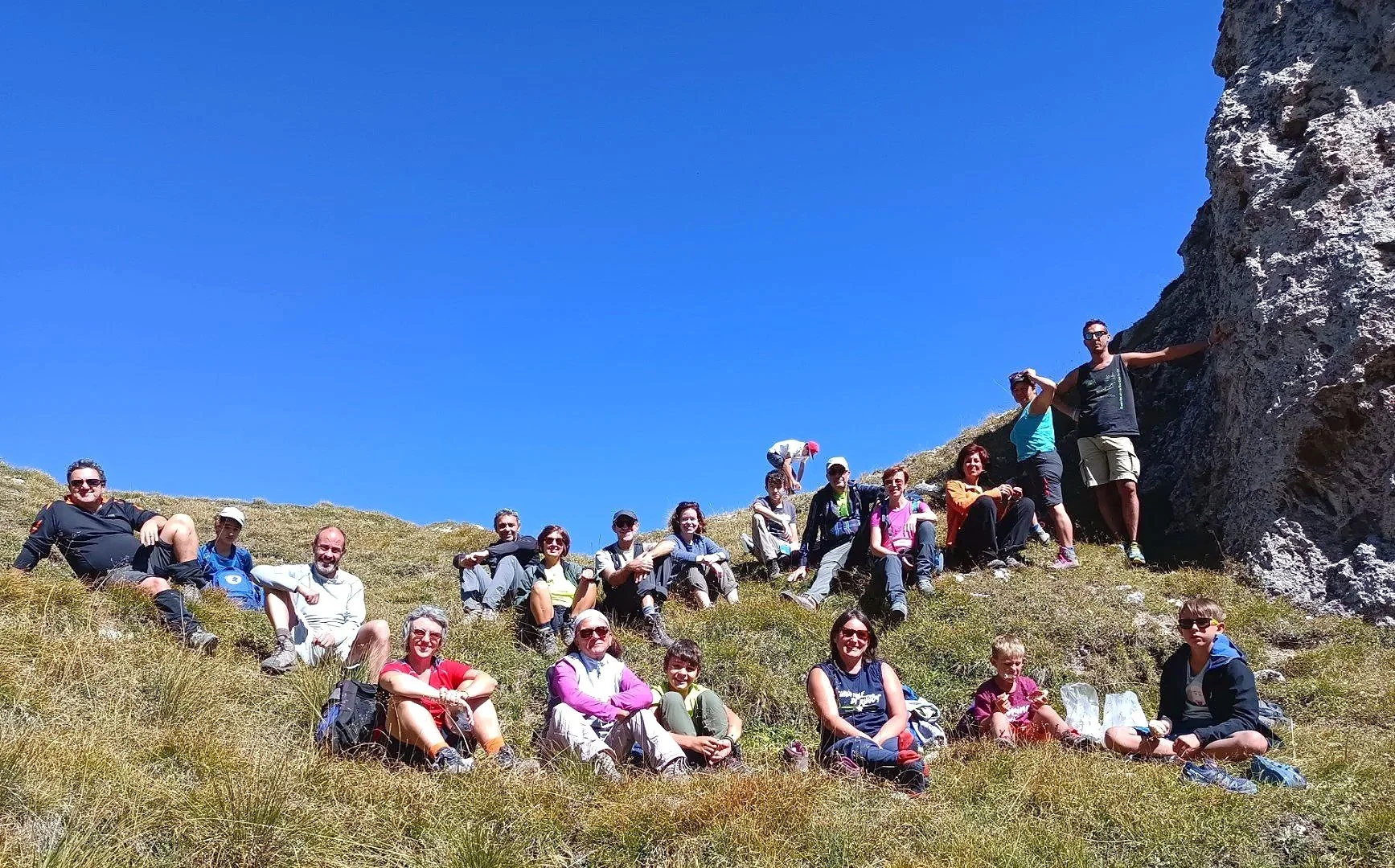 Gruppo di persone sedute e in piedi su un pendio erboso in montagna sotto un cielo blu di fronte a una parete rocciosa.