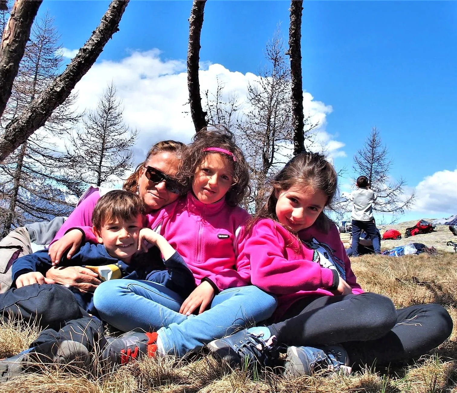 Gruppo di quattro persone, una donna e tre bambini, seduti sull'erba durante una gita in montagna, con alberi spogli e un cielo blu con nuvole sullo sfondo.