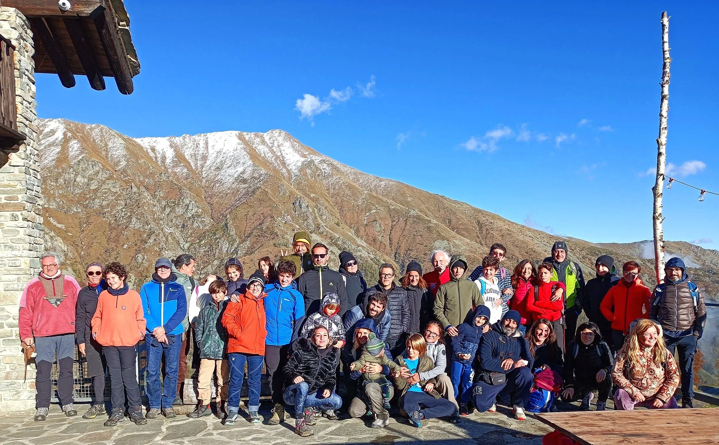 Gruppo di persone di diverse età con abbigliamento invernale, in piedi e sedute, con montagne e cielo azzurro sullo sfondo.