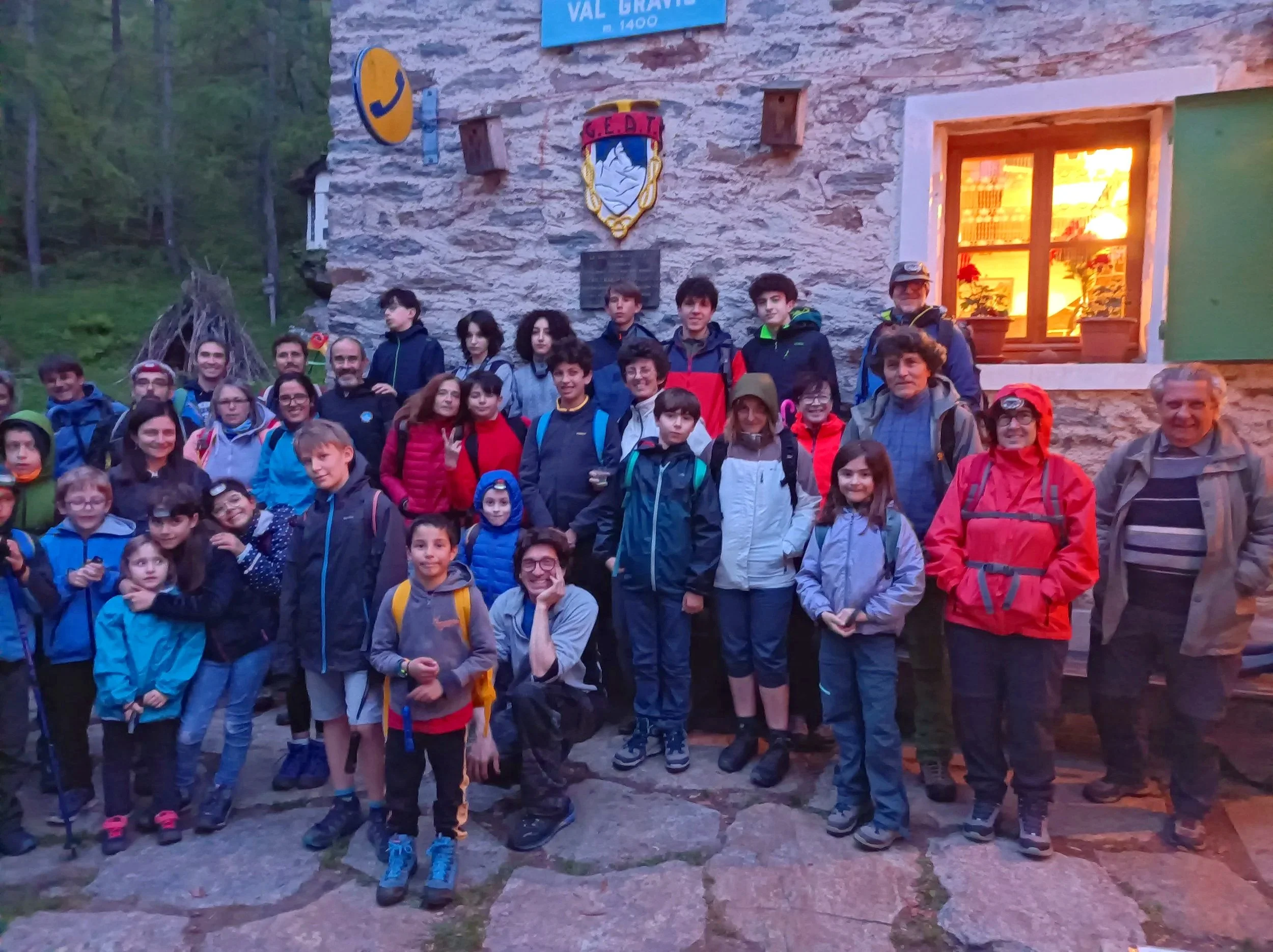 Gruppo di persone, adulti e bambini, davanti a una casa in pietra con una finestra illuminata e un simbolo sulla parete. Sembrano essere in un'escursione o in un'escursione nella natura.