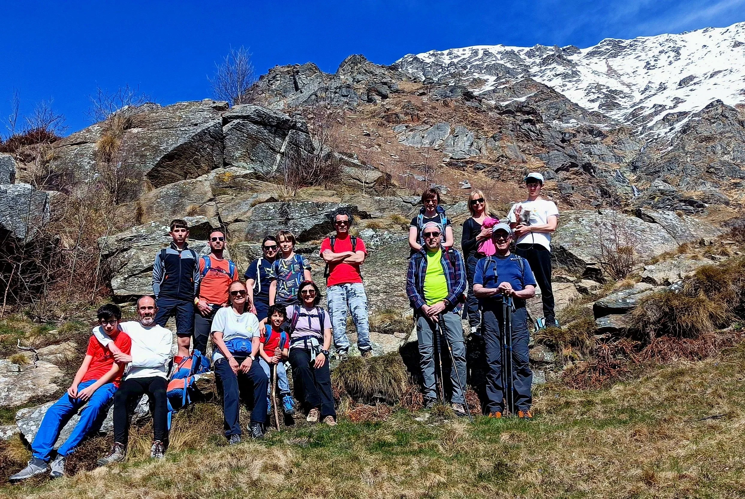 Gruppo di escursionisti con bambini e adulti in montagna, su un terreno erboso con rocce e una montagna innevata sullo sfondo, in una giornata di sole.