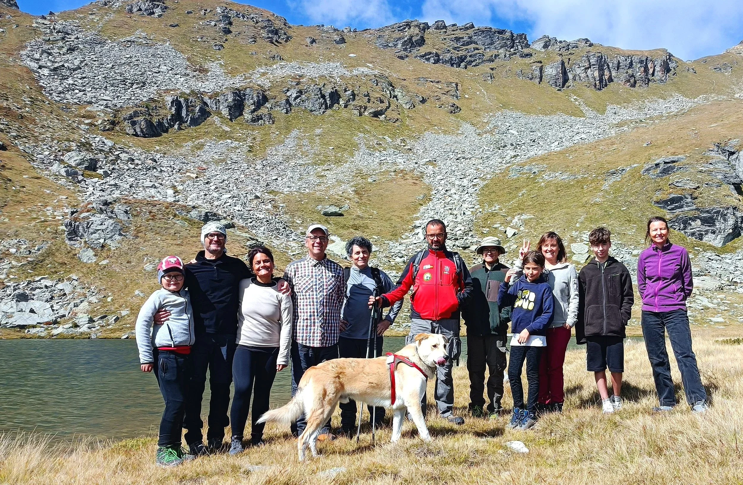 Un gruppo di quattordici persone e un cane da guida si trovano vicino a un lago di montagna con uno sfondo di montagne rocciose e cielo blu, durante un'escursione all'aperto in un paesaggio naturale.