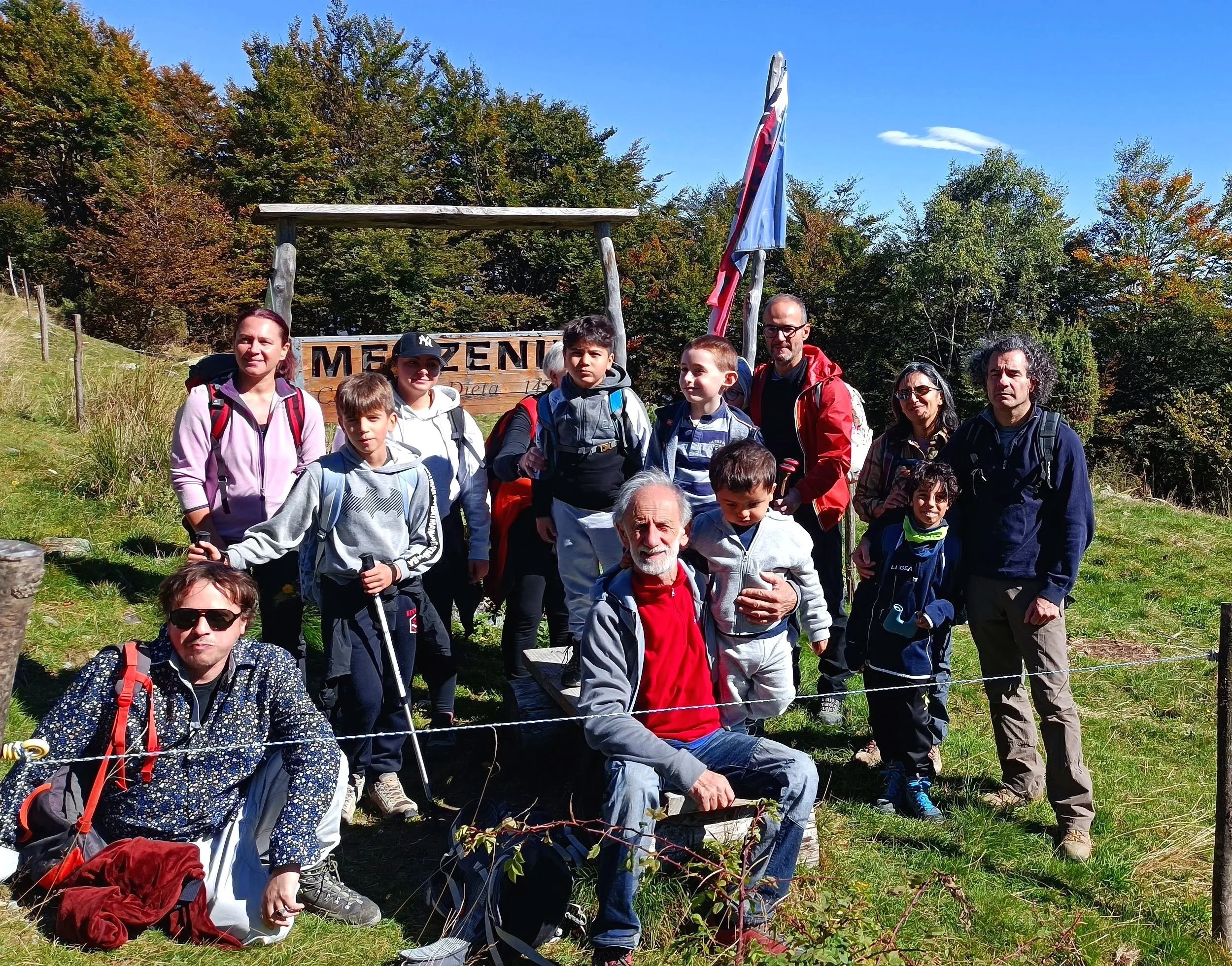 Gruppo di persone, adulti e bambini, in escursione in montagna davanti a un cartello che dice 'MORNICO' con sfondo di natura autunnale, alberi e cielo blu.