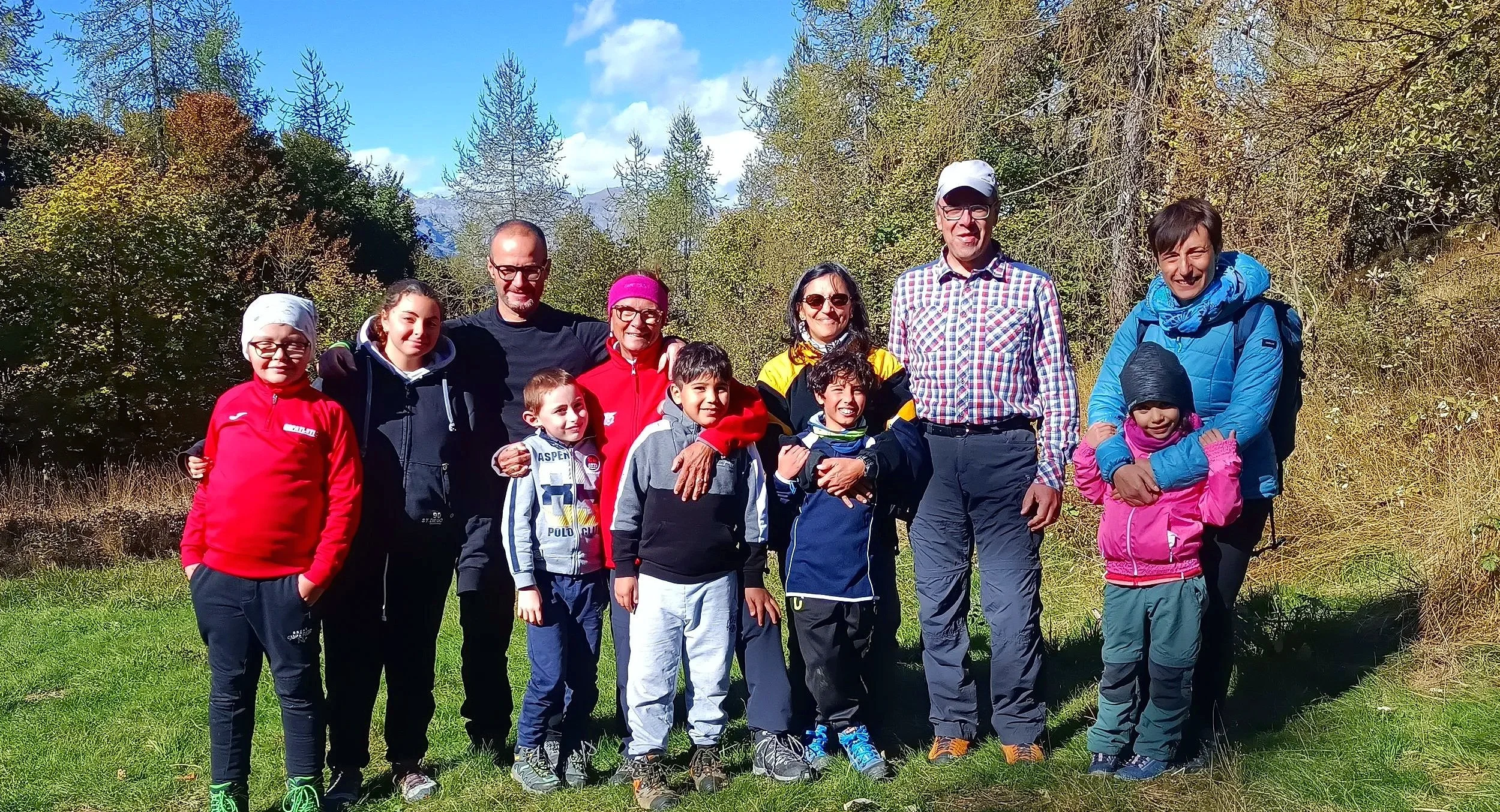Gruppo di persone, adulti e bambini, in un'escursione in natura, con alberi e cielo azzurro sullo sfondo.