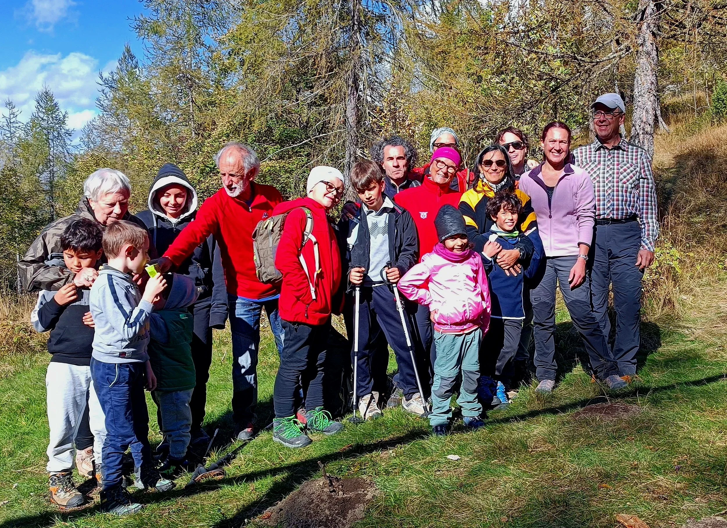 Gruppo di persone di diverse età in escursione in un bosco con alberi e cielo blu.