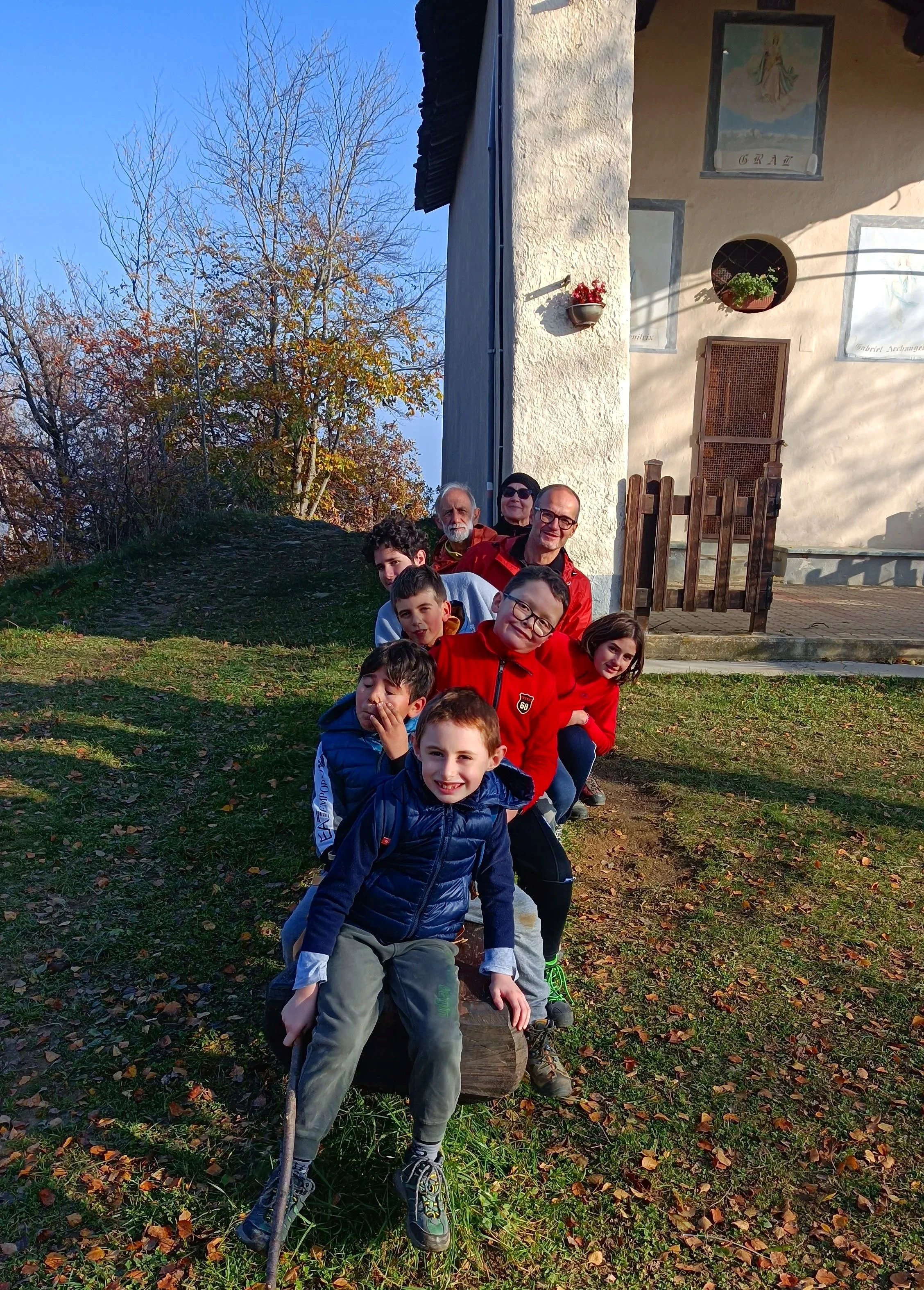 Gruppo di persone, adulti e bambini, in posa all'aperto davanti a una casa, disposta in una fila con alcuni seduti su un tronco e altri in piedi dietro, in un ambiente naturale con alberi e foglie cadute.