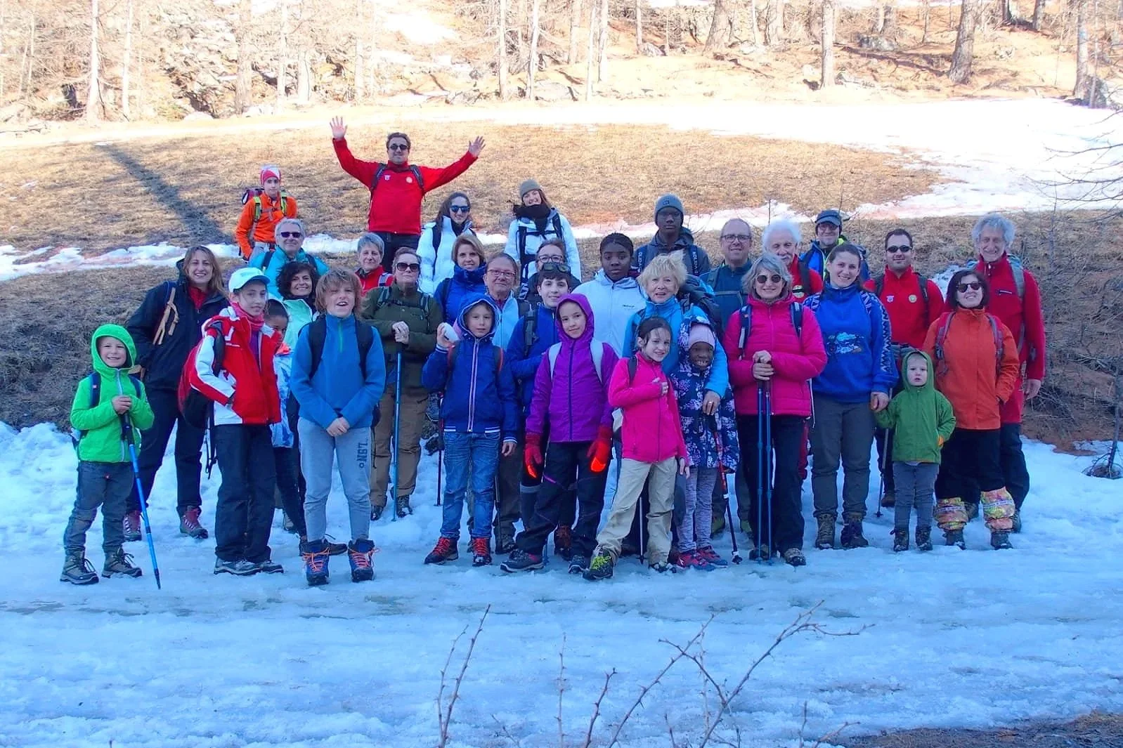 Gruppo di persone di diverse età in escursione in montagna con neve, alcuni con bastoncini, abbigliamento caldo e zaini, in un paesaggio naturale con alberi e terreno innevato.