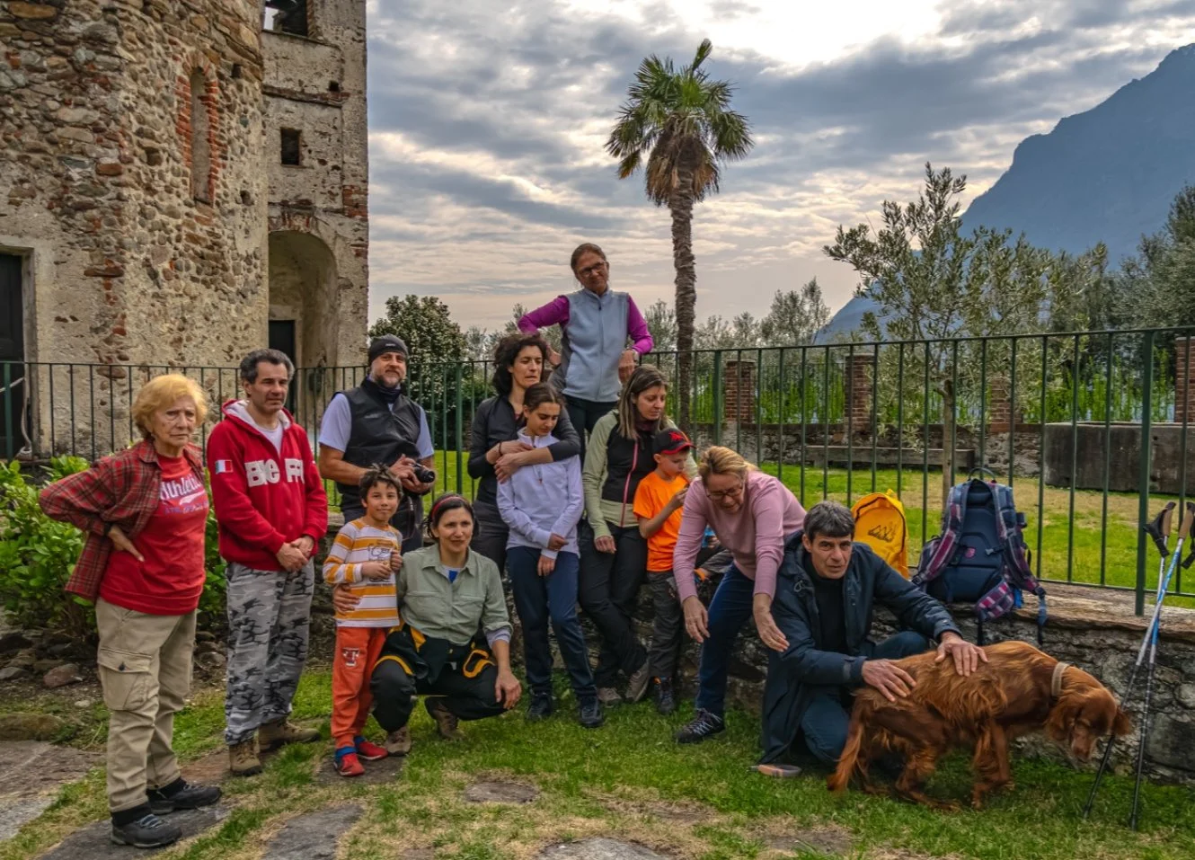 Gruppo di persone di diverse età posano davanti a un'antica struttura di pietra, con alcuni membri che interagiscono con un cane, in un setting naturale con alberi, una palma e montagne sullo sfondo, su un prato verde.