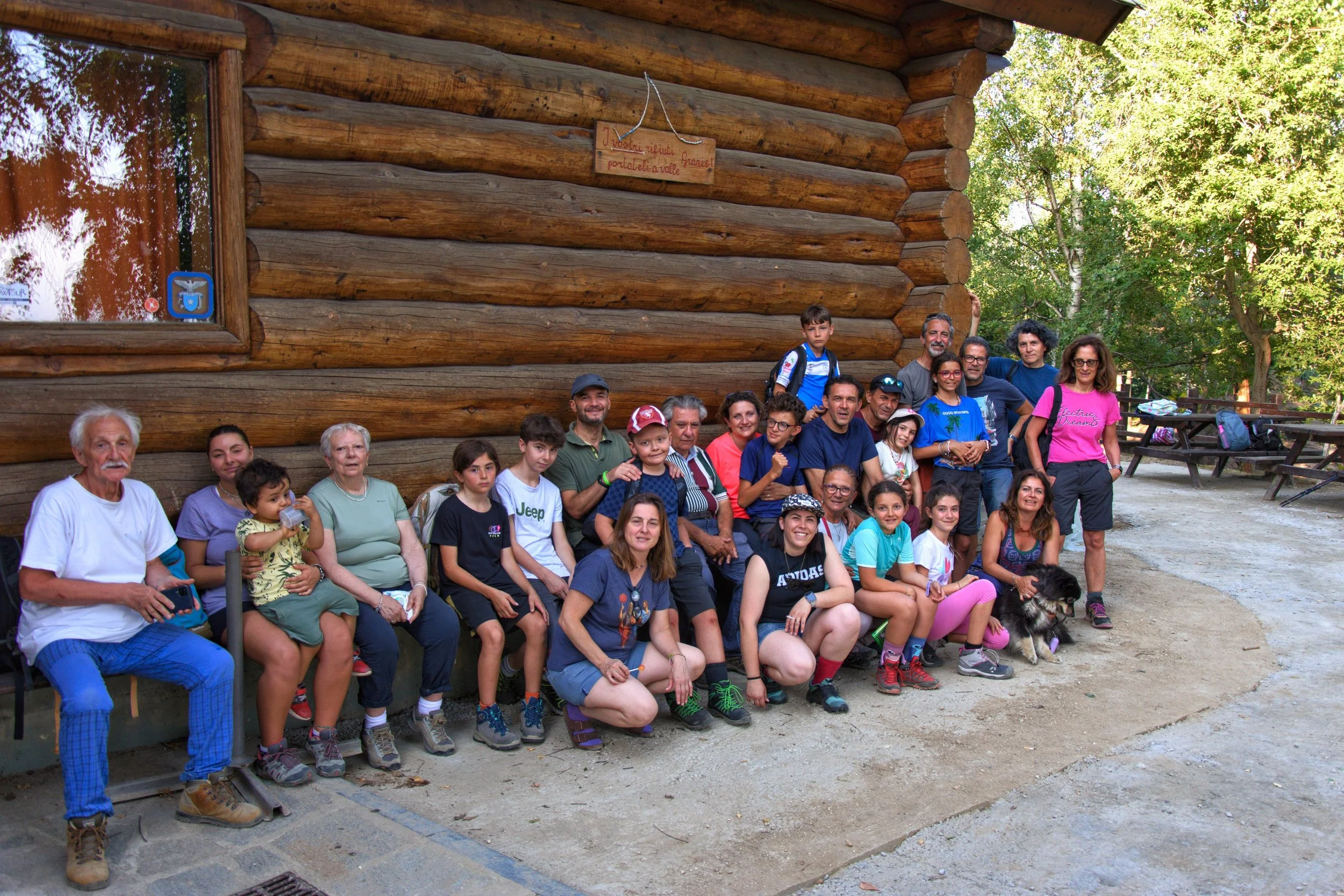 Gruppo di persone di diverse età che posano davanti a una baita in legno, alcune sedute su una panchina e altre in piedi, in un'area naturale con alberi.