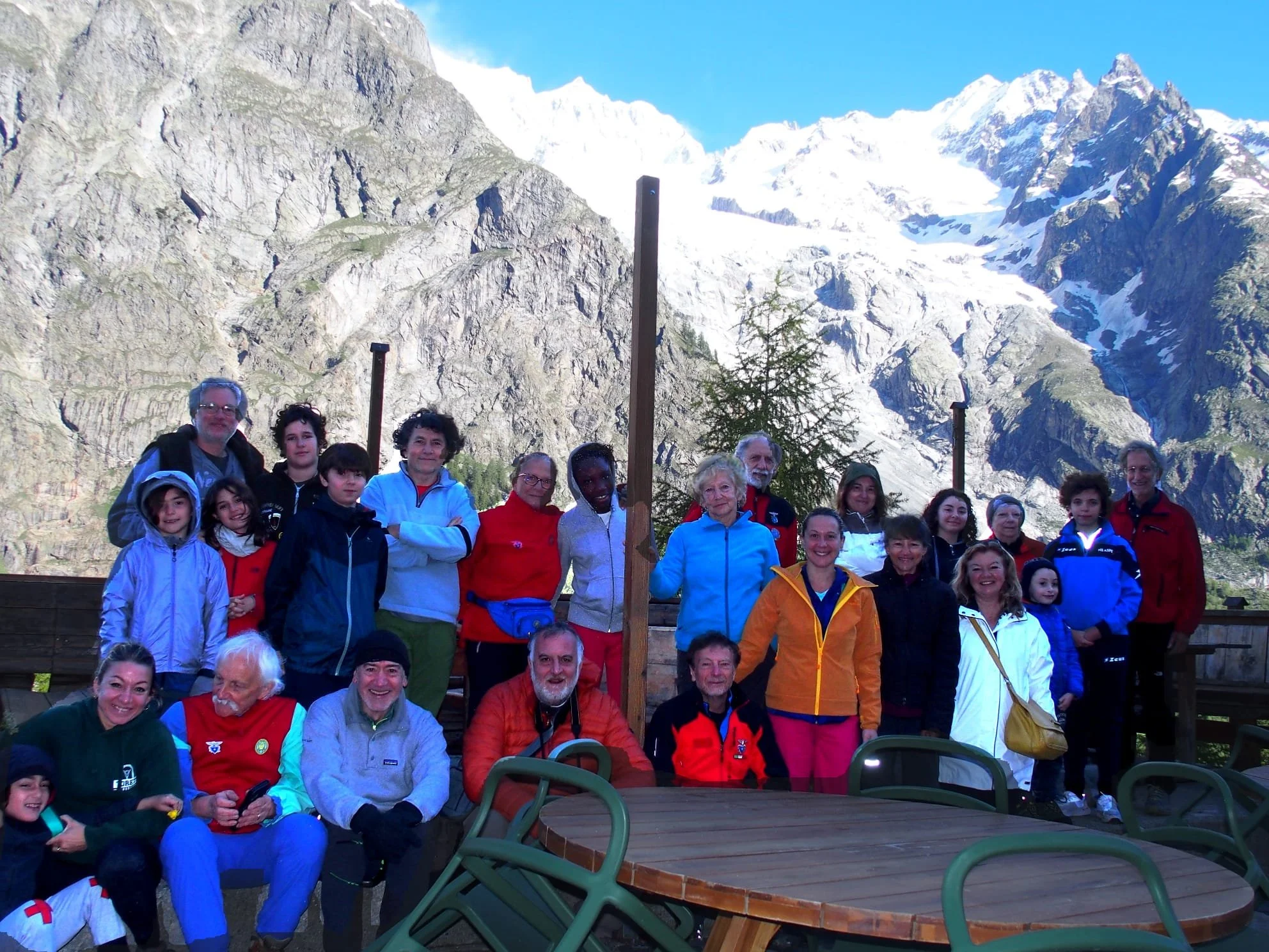 Un gruppo di persone davanti a montagne con neve, alcune sedute e altre in piedi, con sfondo un paesaggio montano.