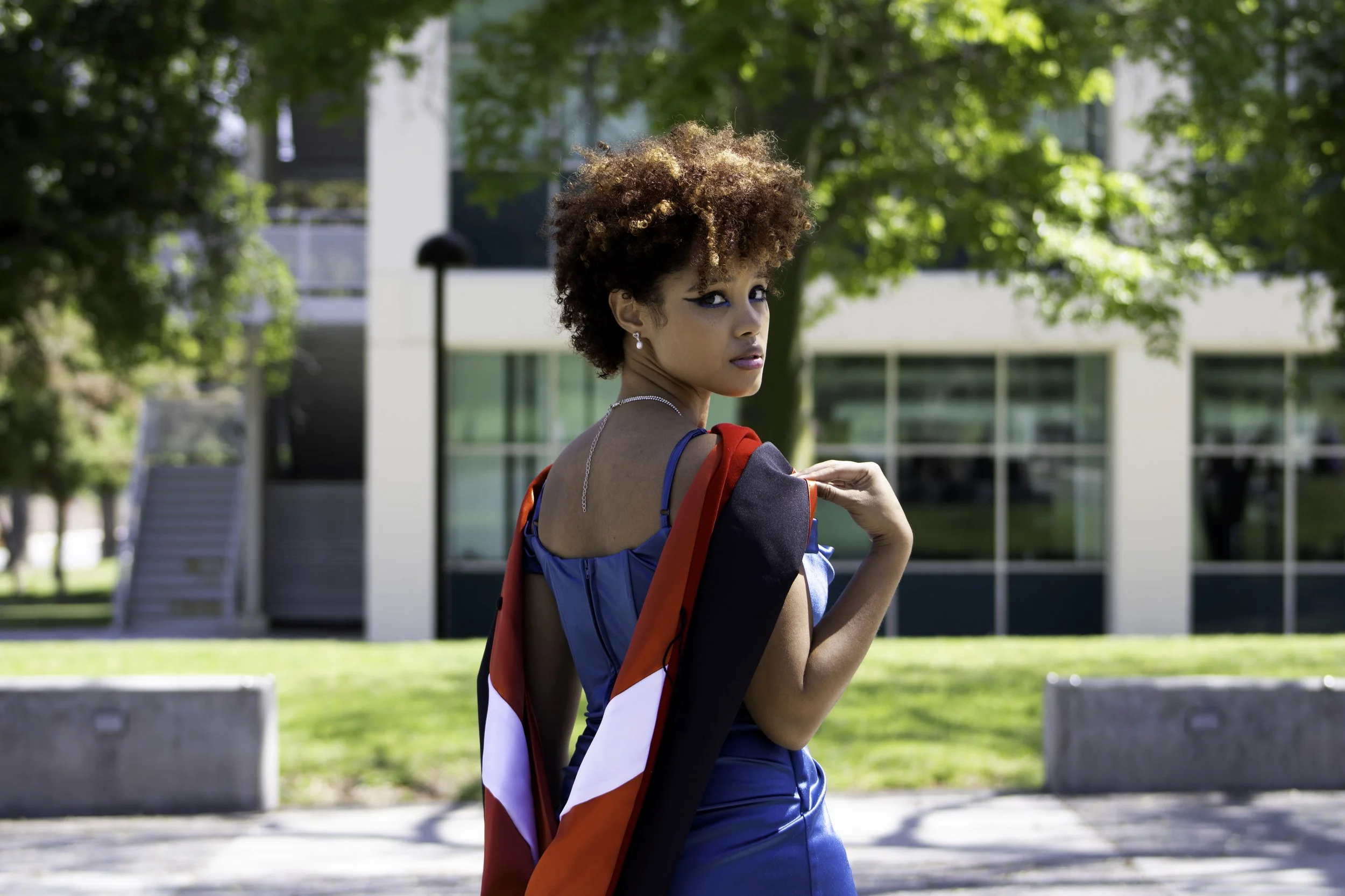 Eurice standing outdoors with trees and a modern building in the background. She is wearing a blue dress and has a red, white, and black sash draped over her shoulder, while looking over her shoulder at the camera.