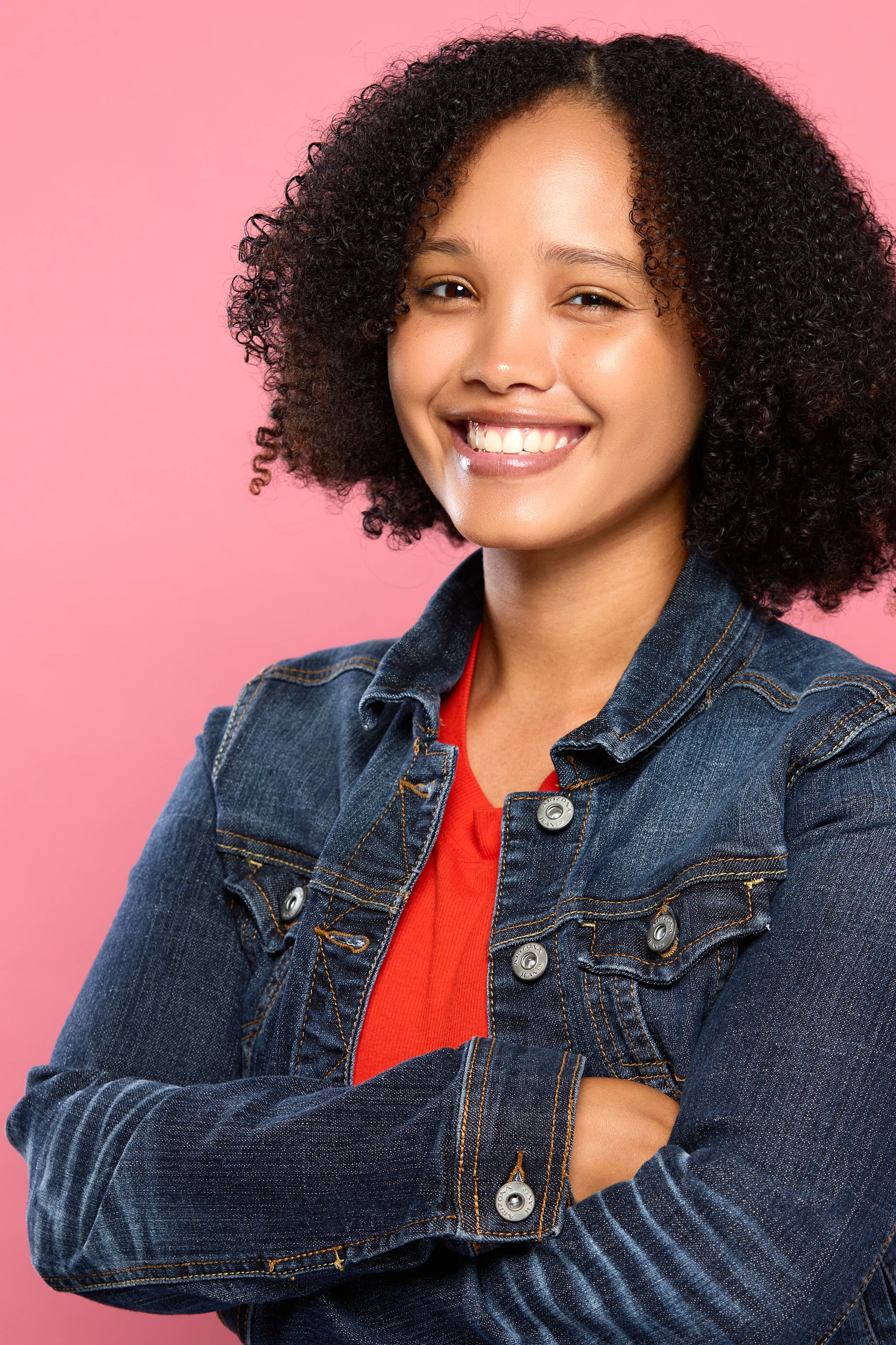 Eurice, wearing a denim jacket over a red shirt, standing against a pink background.