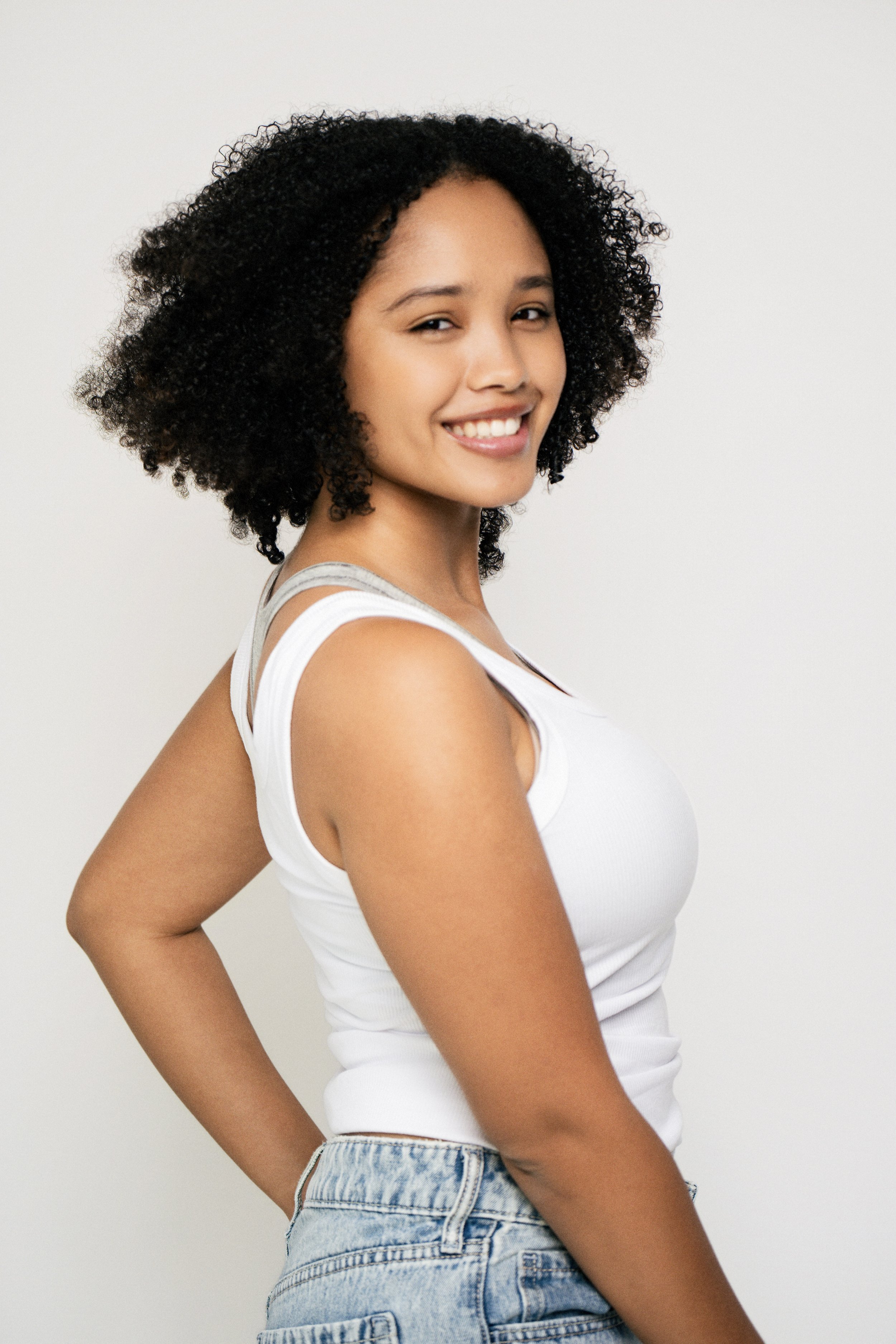 Eurice smiling, wearing a white tank top and jeans, standing against a plain background.