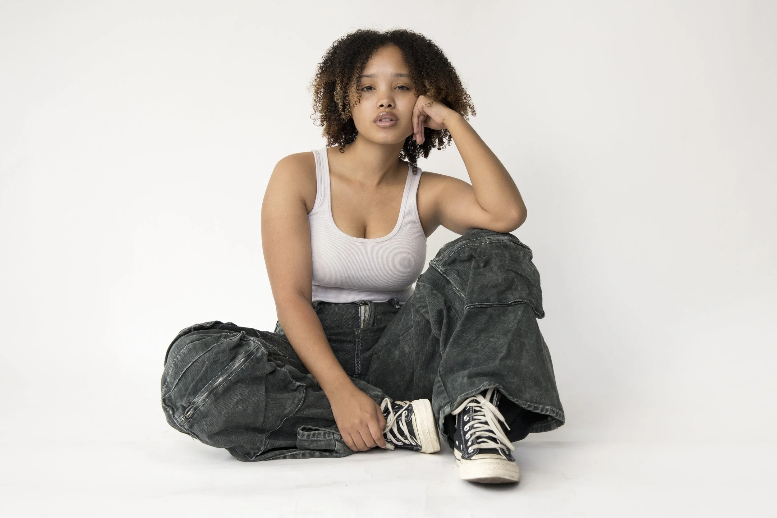 Eurice sitting on the floor, wearing a white tank top, baggy black jeans, and black sneakers, against a plain white background.