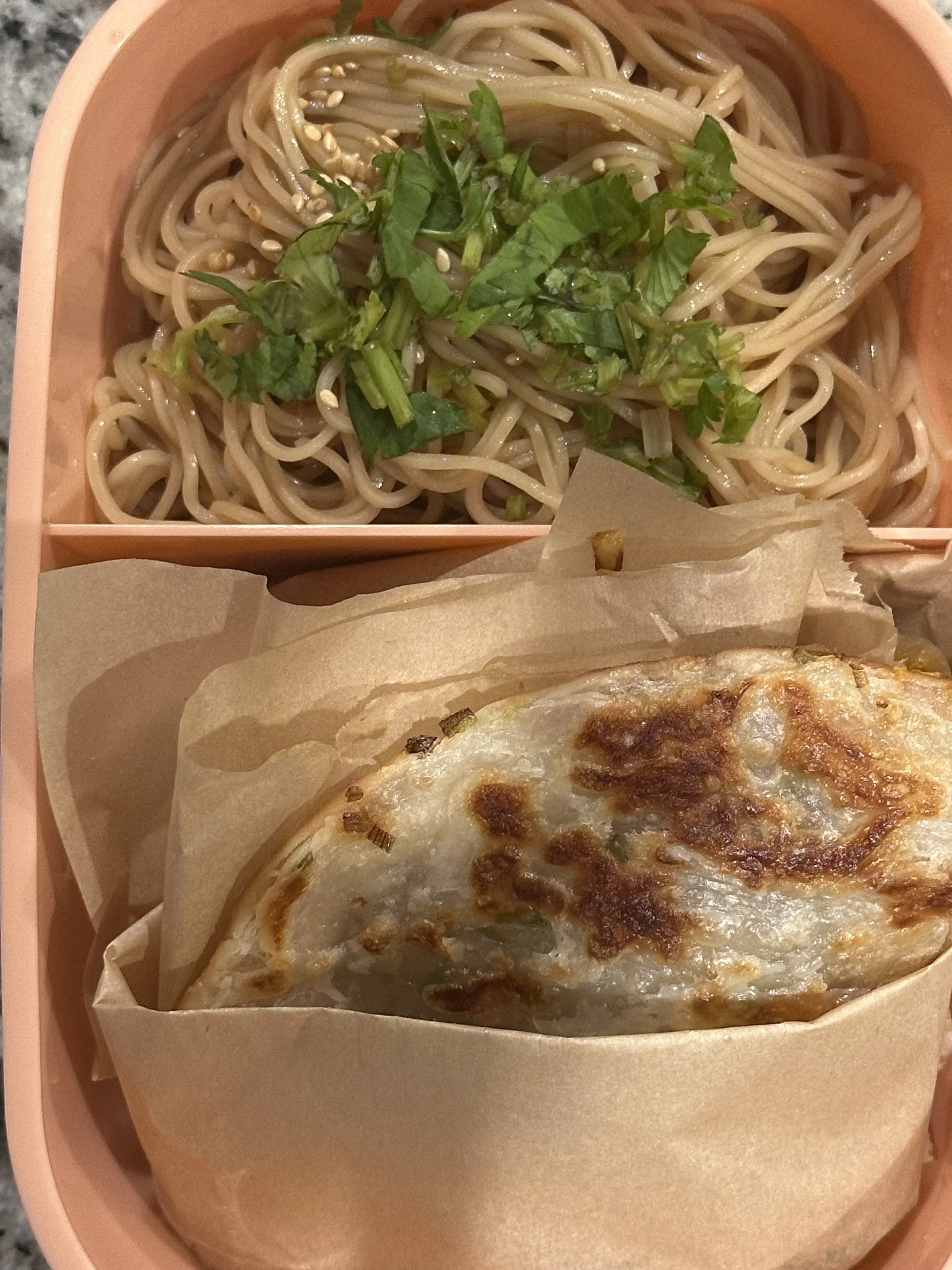 A pink bento box contains a noodle salad garnished with chopped cilantro and sesame seeds, and a grilled cheese sandwich wrapped in parchment paper.