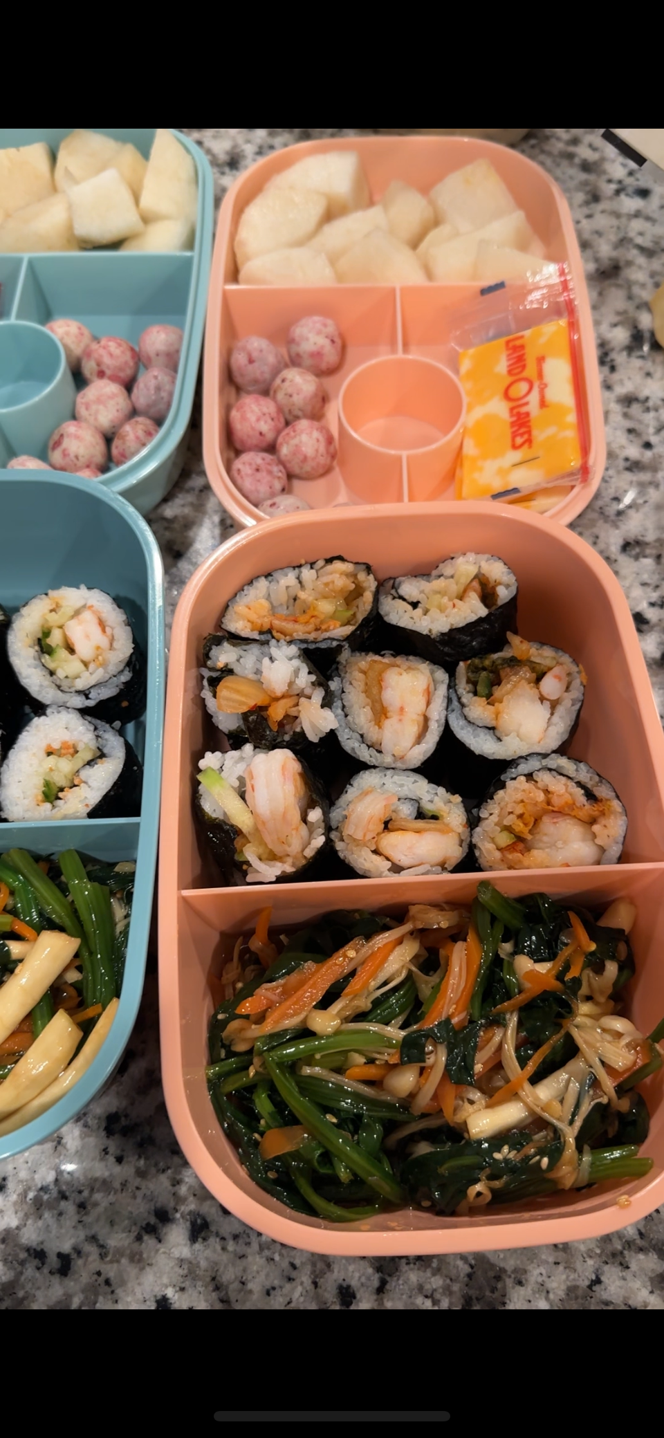 A colorful bento box filled with sushi rolls, mixed vegetables, and fruit. The sushi rolls contain shrimp and vegetables wrapped in seaweed, and the vegetables include green beans, carrots, and music, in separate compartments. The fruit section contains diced cantaloupe, honeydew, and small fruit balls.