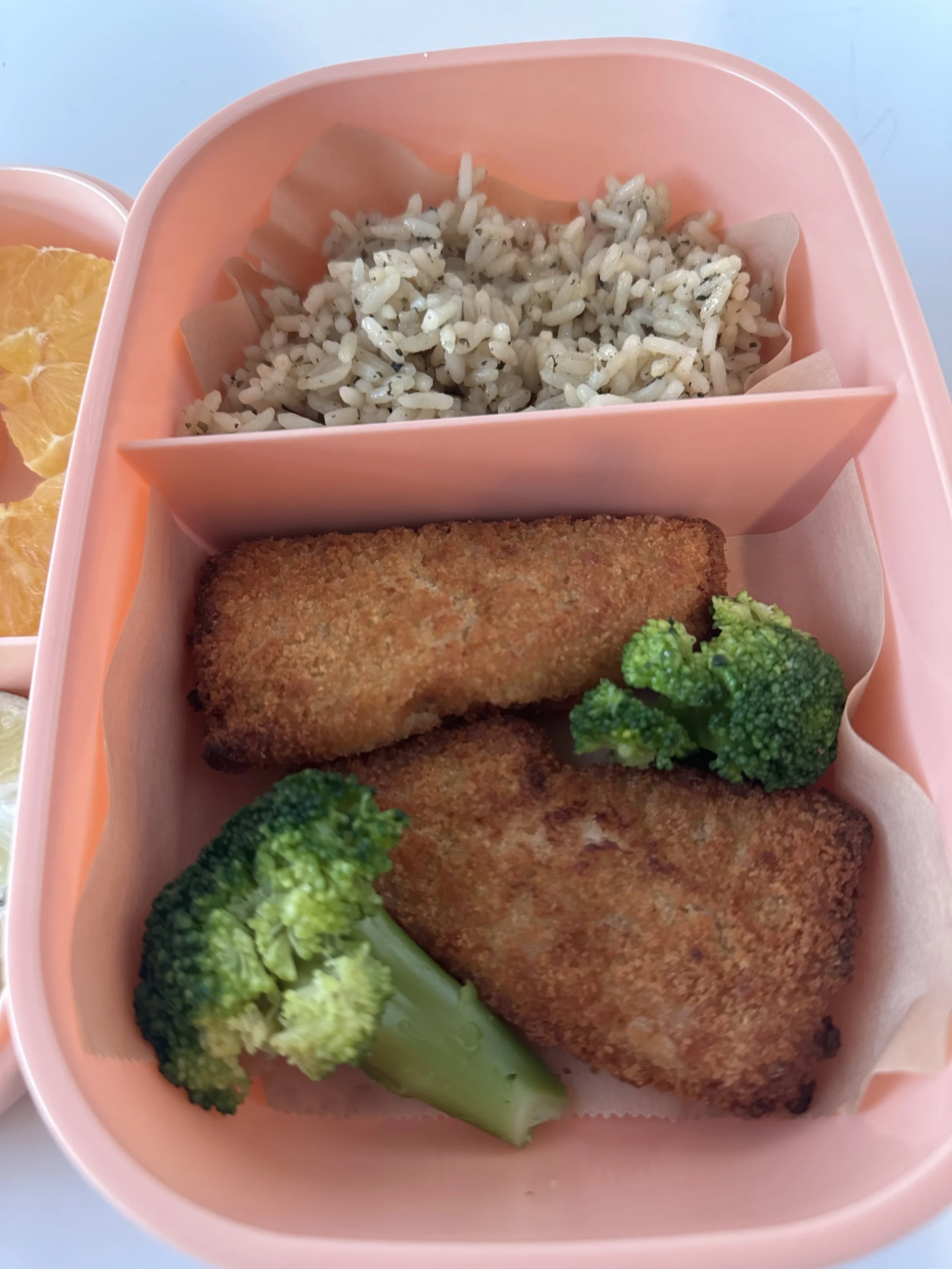 A pink bento box with two pieces of breaded fried fish, broccoli florets, a serving of rice with black pepper, and a side of orange slices.