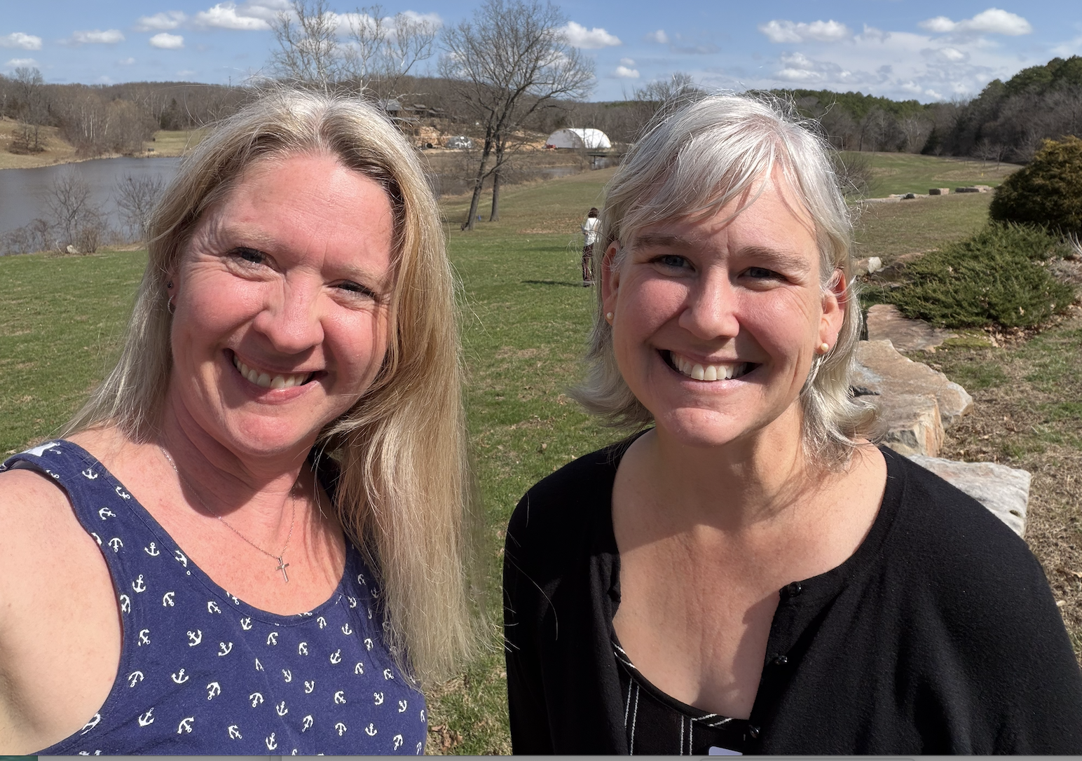 Two women smiling outdoors near a lake with trees and sky in the background.