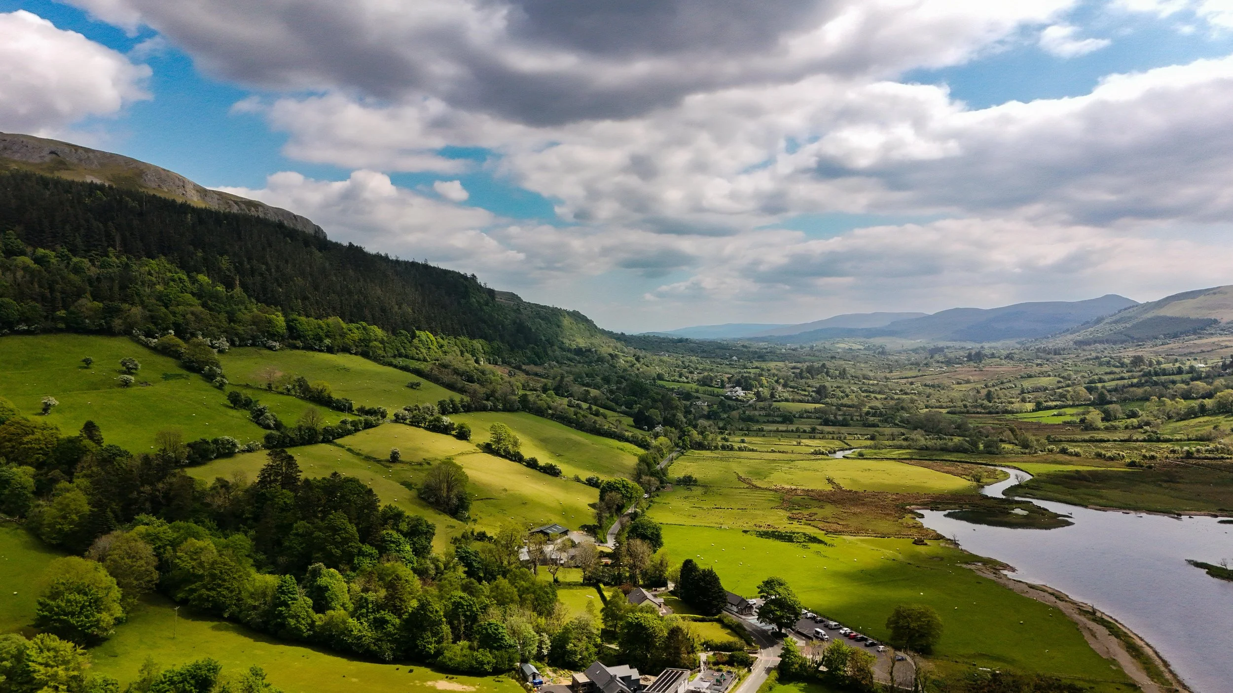 A scenic view of lush green fields, trees, a river, and rolling hills under a partly cloudy sky.
