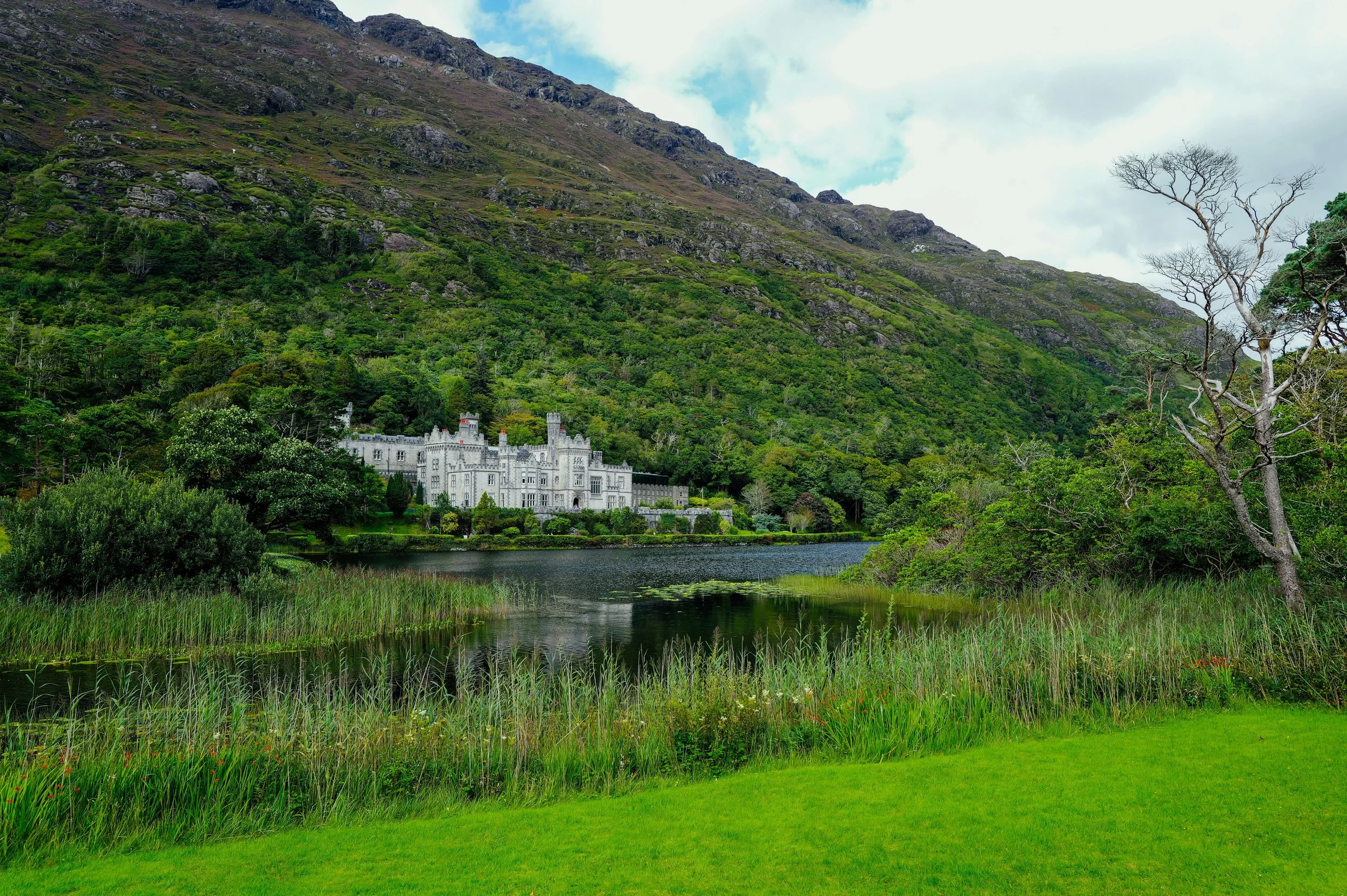 A large white castle sits near a lake, surrounded by lush green trees and hills with rocky slopes, under a partly cloudy sky.