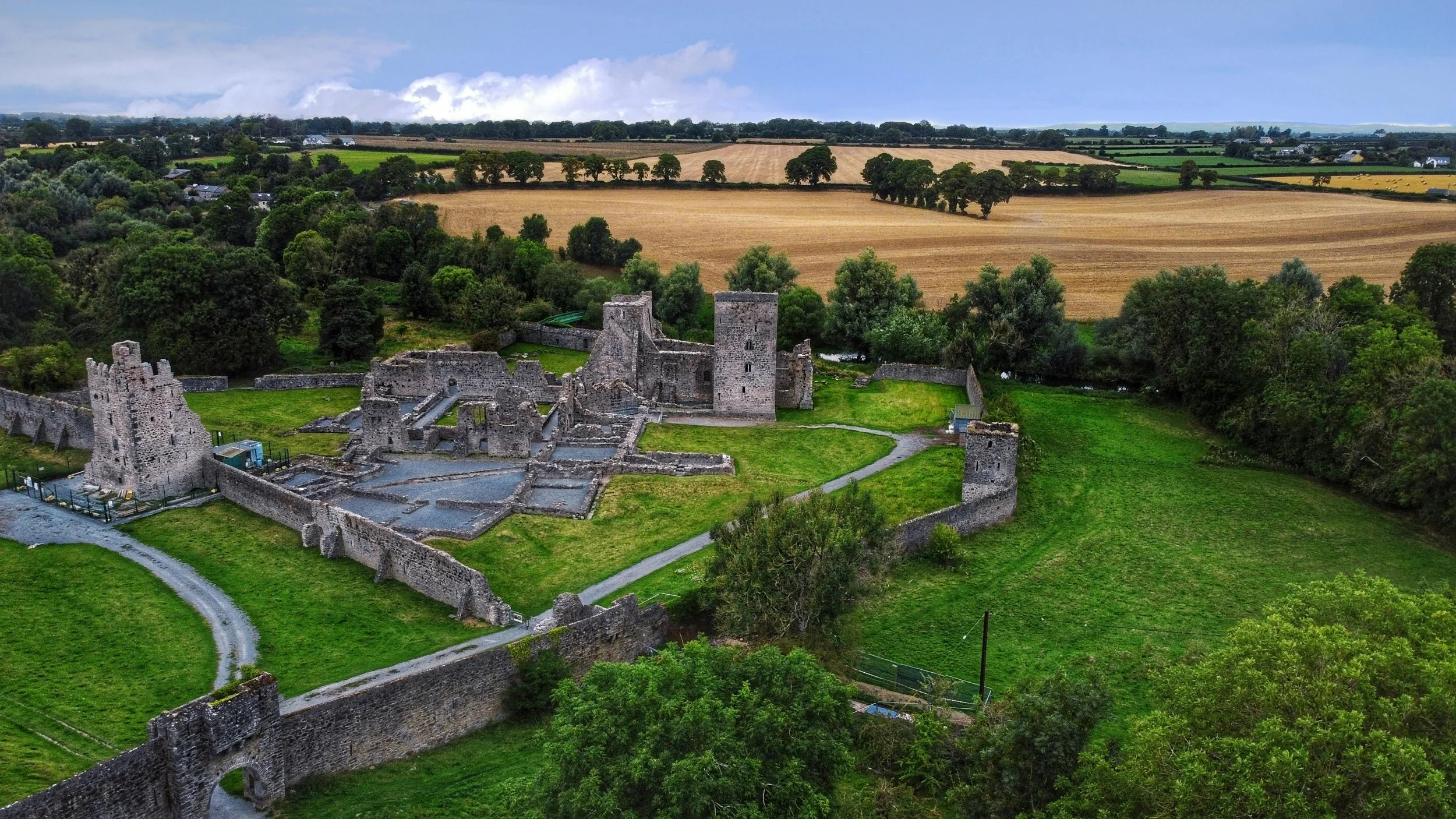 Aerial view of an ancient stone castle surrounded by green grass, trees, and farmland under a cloudy sky.
