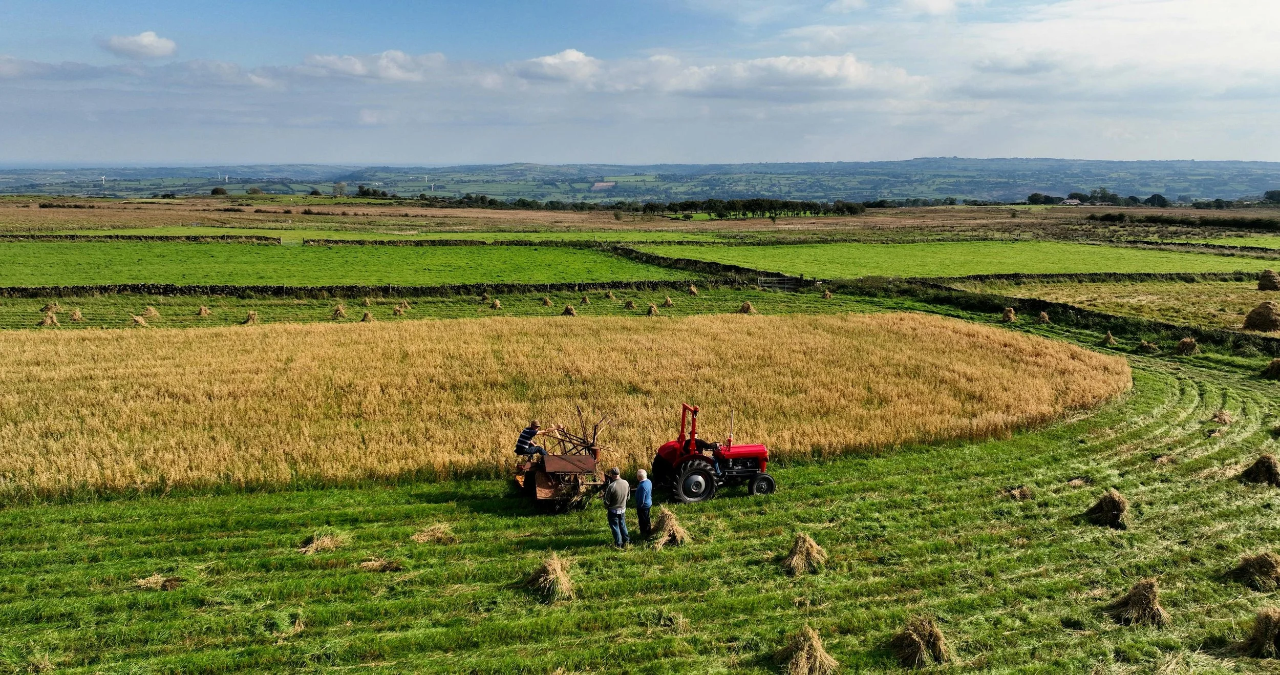 Farmers harvesting crops with a red tractor and a trailer in a lush green landscape with multiple fields and haystacks under a partly cloudy sky.