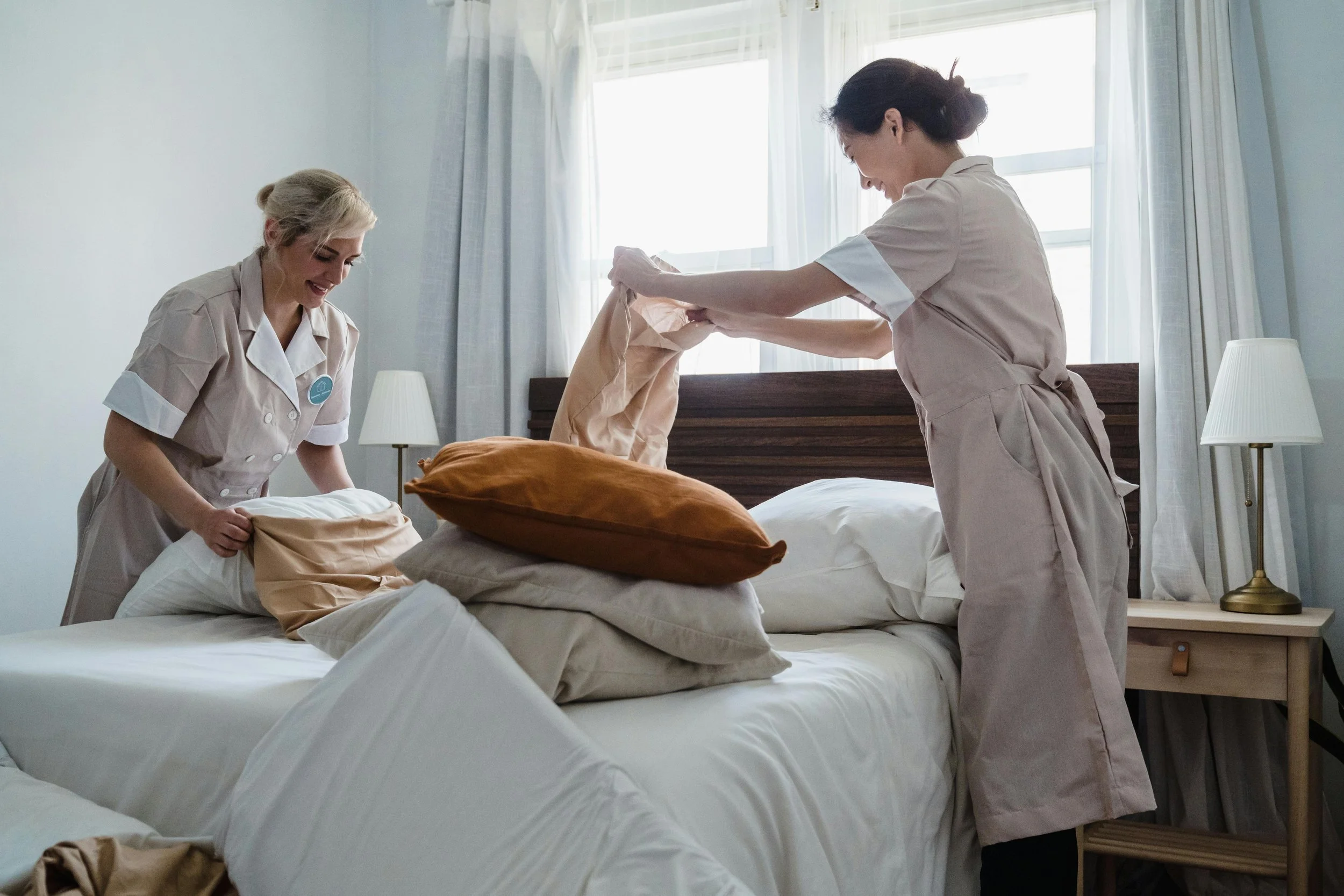 Two housekeepers making a bed together in a well-lit bedroom.