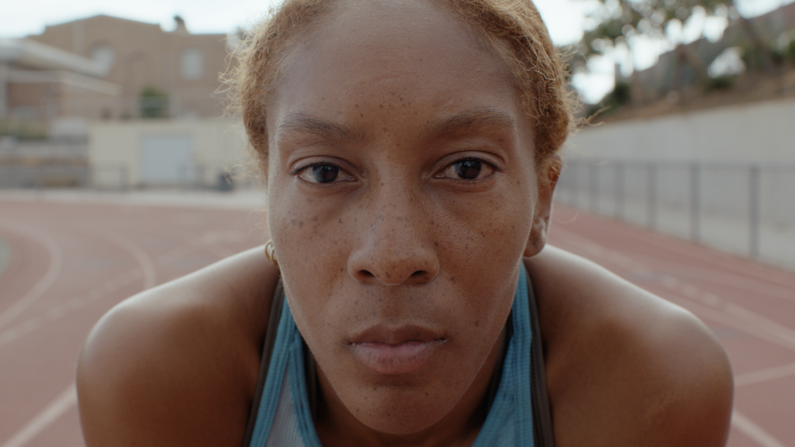 Close-up of a female athlete with freckles, wearing a blue sports top, preparing to race on an outdoor track.