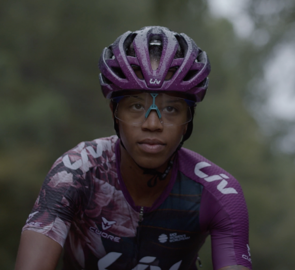 A young cyclist wearing a purple helmet and sports sunglasses, dressed in a purple and patterned cycling jersey, riding outdoors.