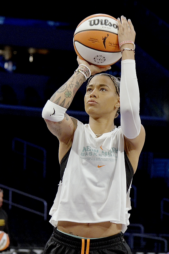 A female basketball player wearing a white sleeveless shirt and black shorts lined with orange, practicing free throws on a basketball court. She is holding a Wilson basketball above her head, preparing to shoot, with tattoos on her left arm and a focused expression.