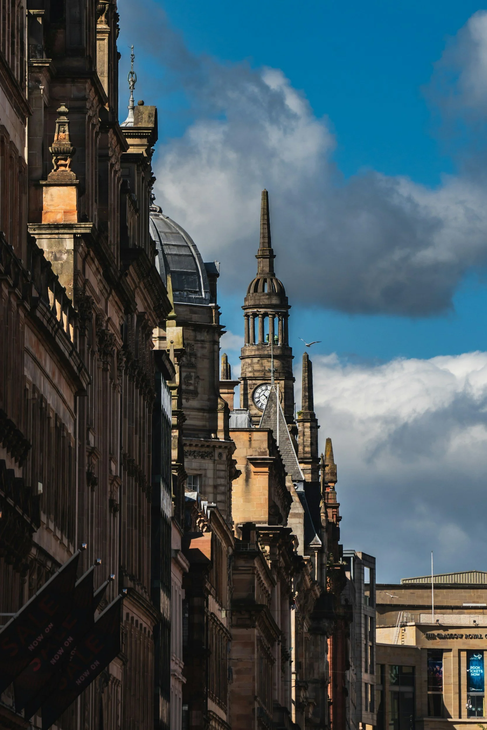 A city street scene with historic buildings featuring ornate architecture, a clock tower, and a spire, under a blue sky with clouds and a seagull flying.