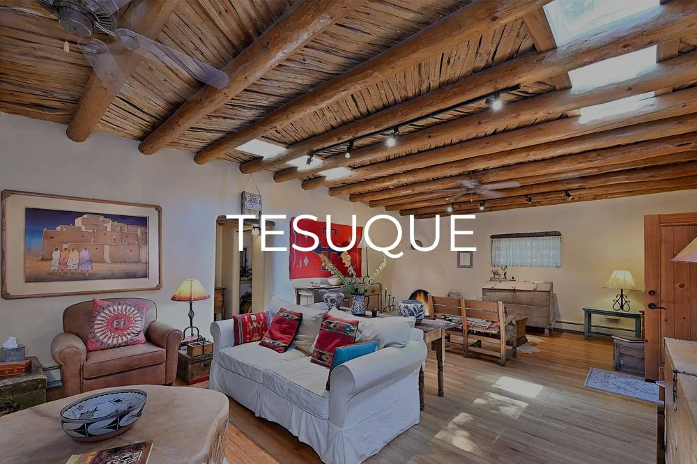 A cozy living room with a rustic wooden ceiling, a ceiling fan, and warm lighting. There is a beige armchair, a white couch with red and black pillows, a wooden dining table, and various furniture pieces and decorations, including paintings and lamps