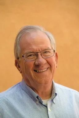 A smiling elderly man with glasses, wearing a blue collared shirt, standing against a beige background.
