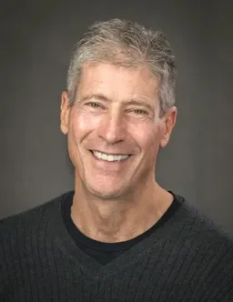 A smiling middle-aged man with gray hair and a black sweater, standing against a dark background.