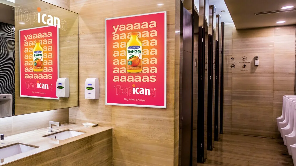 A restroom with a mirror, two hand sanitizer dispensers, and a Tropicana juice advertisement on the wall.