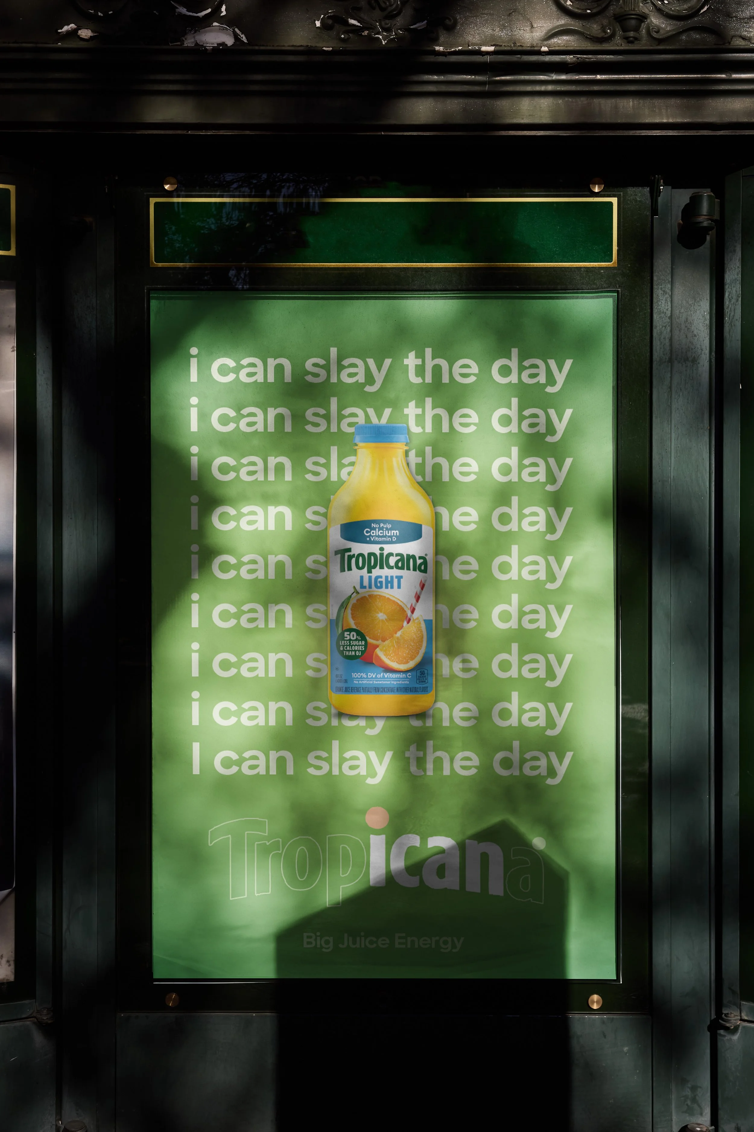 A green advertisement poster for Tropicana Light orange juice, featuring a large image of the juice bottle with a blue cap and a straw in the glass, with the slogan repeating 'I can slay the day' in white text.