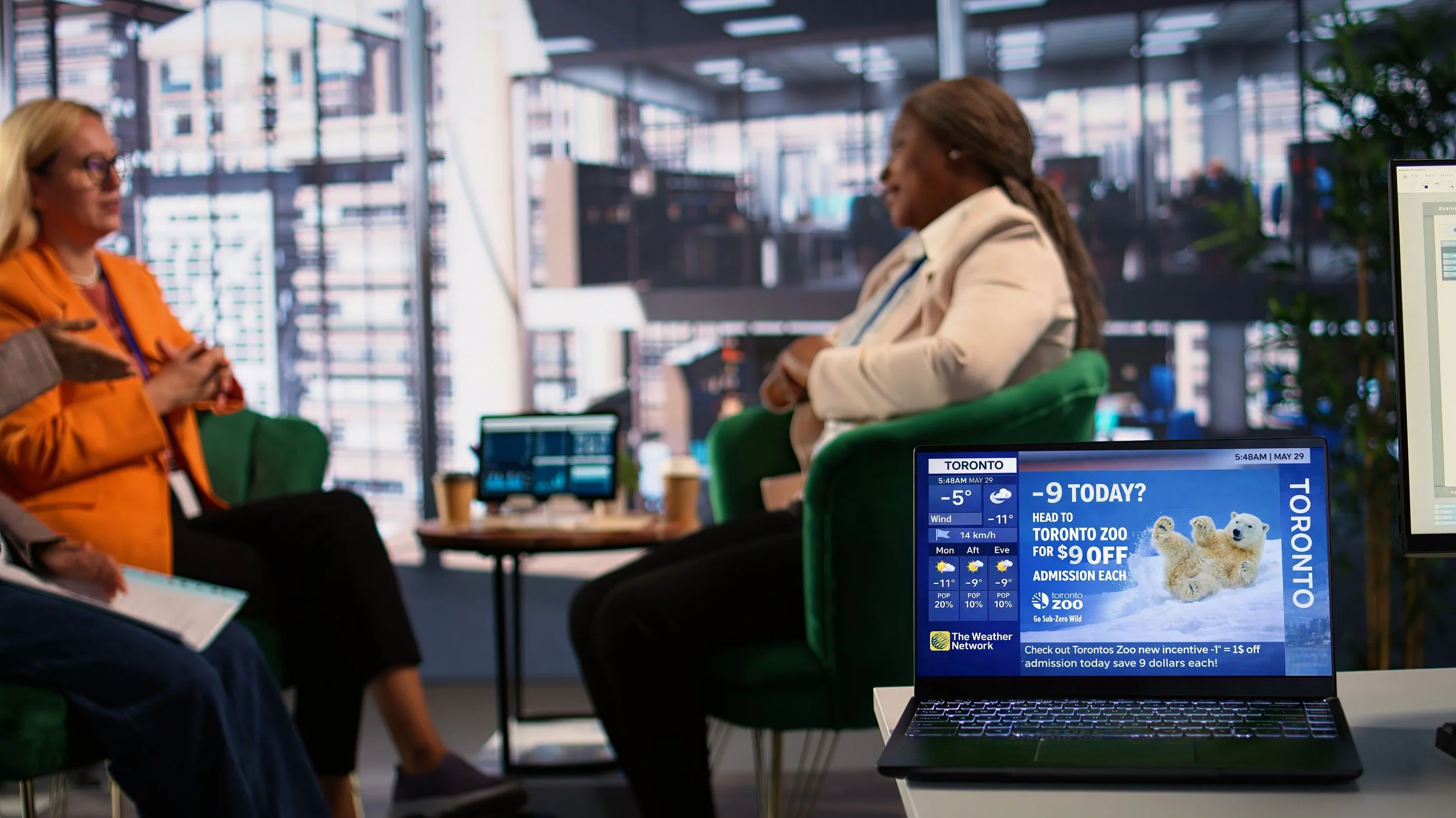 Two women in conversation in a modern office space with large windows. A laptop on a table displays an advertisement about Toronto Zoo with a wildlife image and a weather widget.