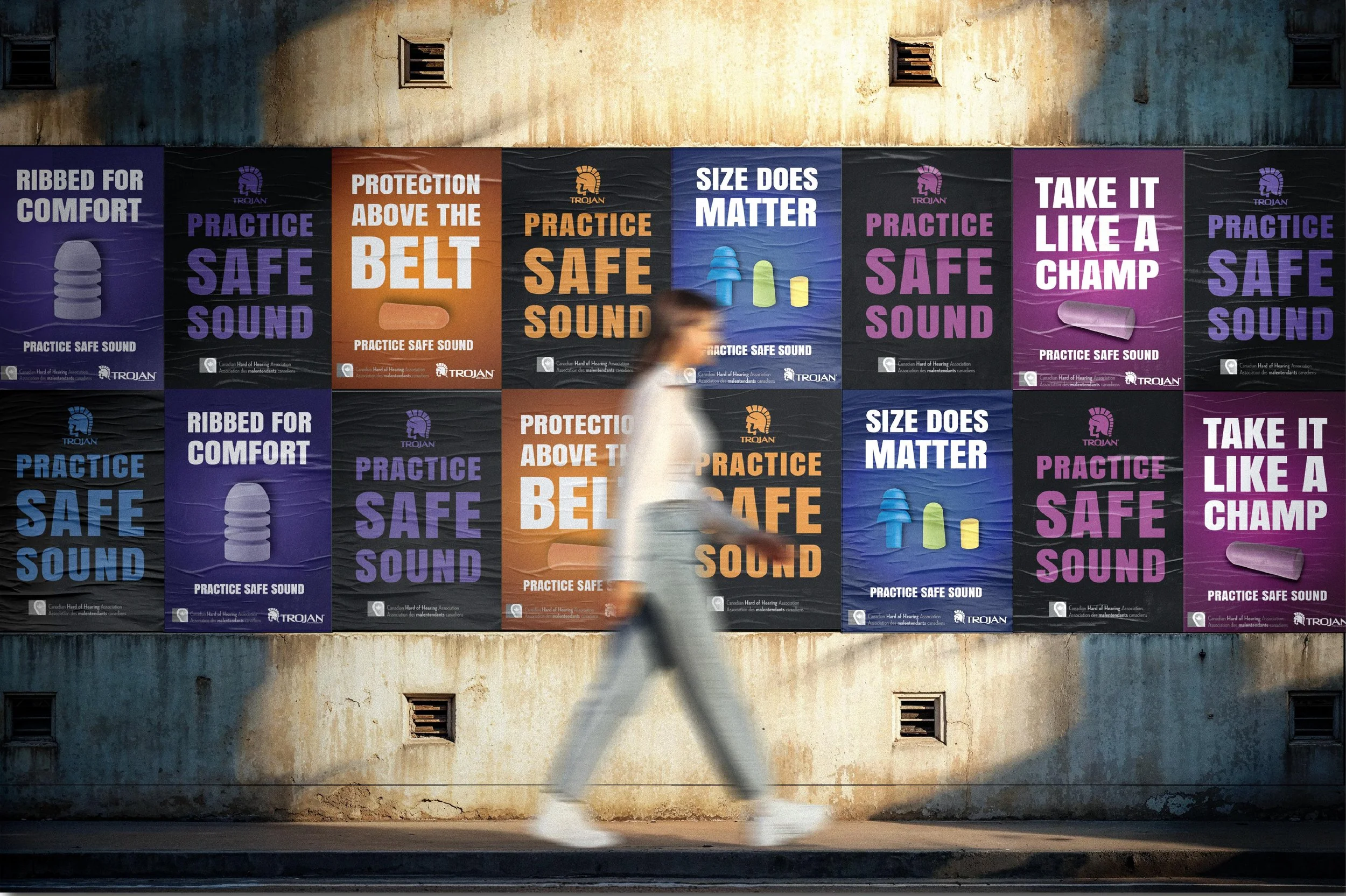 A man walking past a wall covered with colorful posters promoting safe sound practices, wearing a white hoodie and gray pants.